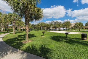 A park with a paved walkway, palm trees, and a building in the background.