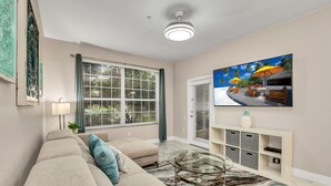 En moderne stue med en beige sofa, et glasbord og et billede af en tropisk strand.