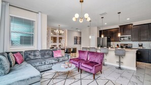 Ein modernes Wohnzimmer mit einem grauen Ecksofa, einem runden Couchtisch und einem violetten Sessel. Im Hintergrund ist eine Küche mit dunklen Schränken und Edelstahlgeräten zu sehen.