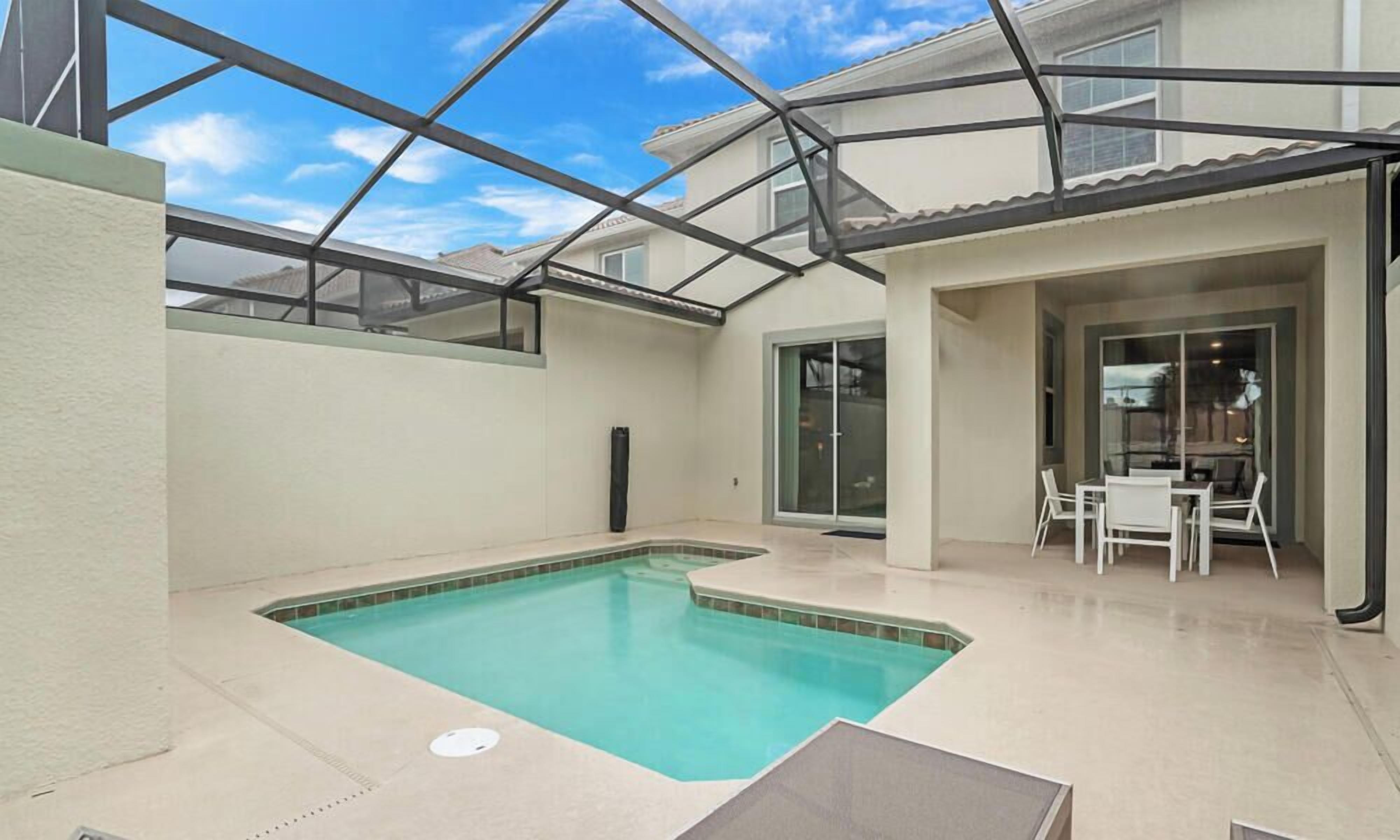 Una piscina con techo de vidrio, un área de patio con mesa y sillas, y un ventanal corredero que da a un espacio interior.