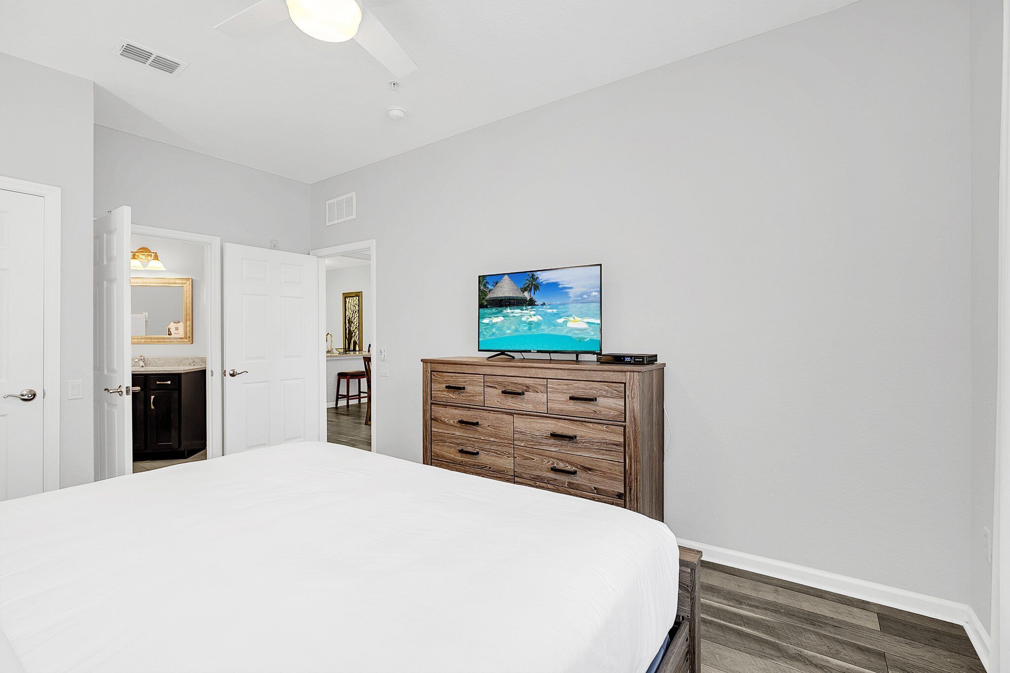 Un dormitorio con cama, cómoda de madera, televisor sobre la cómoda y una puerta que da a otra habitación.