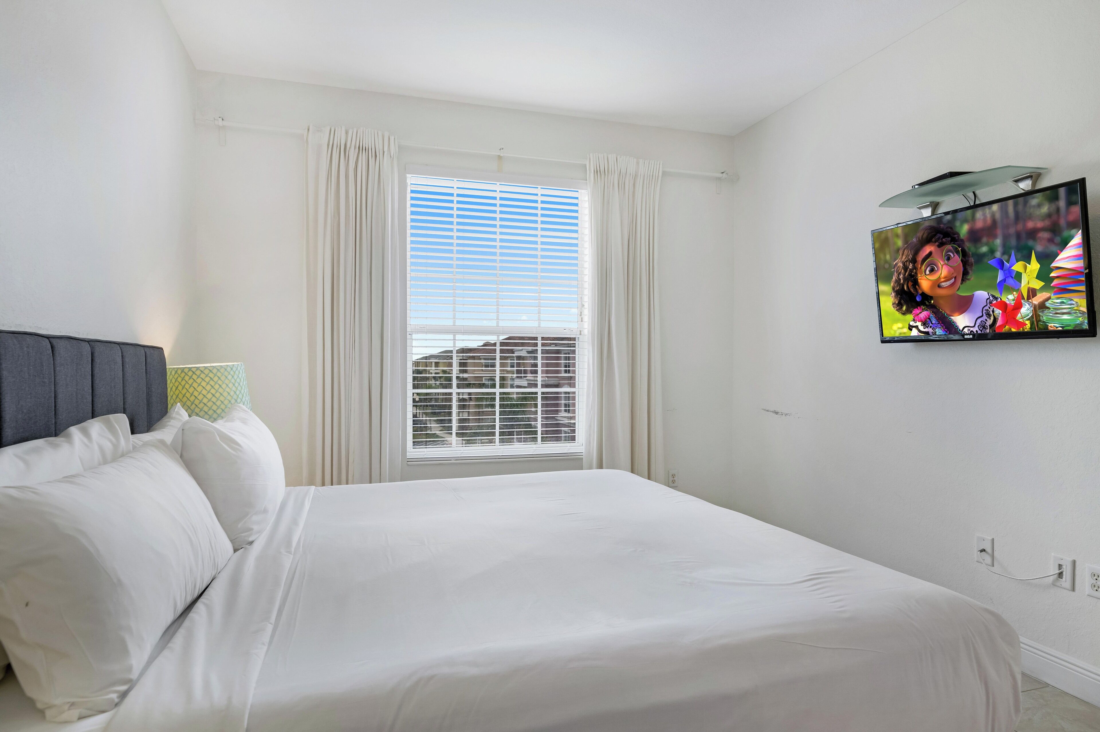 Een slaapkamer met een bed, gordijnen, een raam met zonwering en een aan de muur bevestigde televisie.