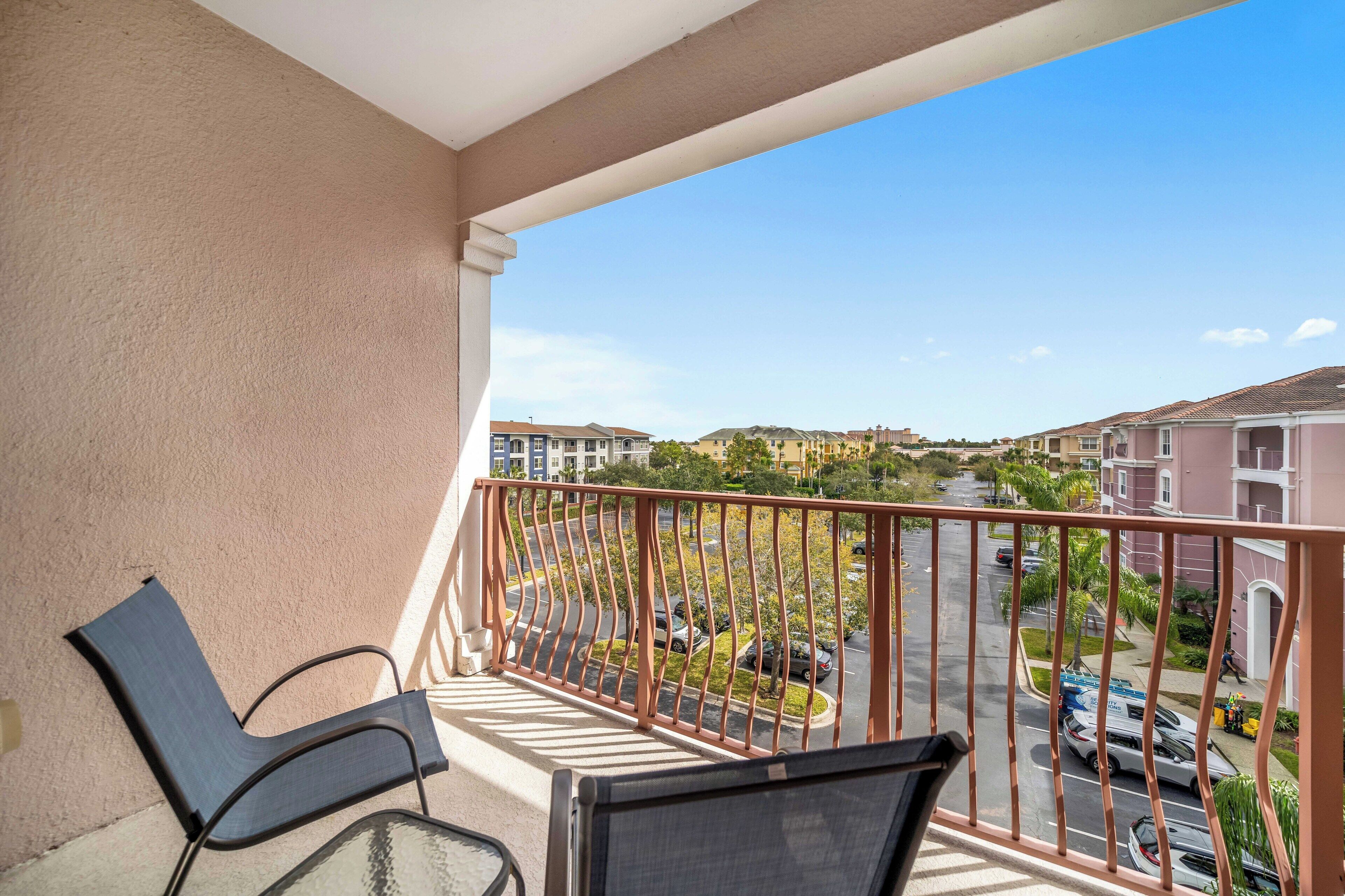 A balcony with two chairs and a glass table, overlooking a residential area with houses and a road.