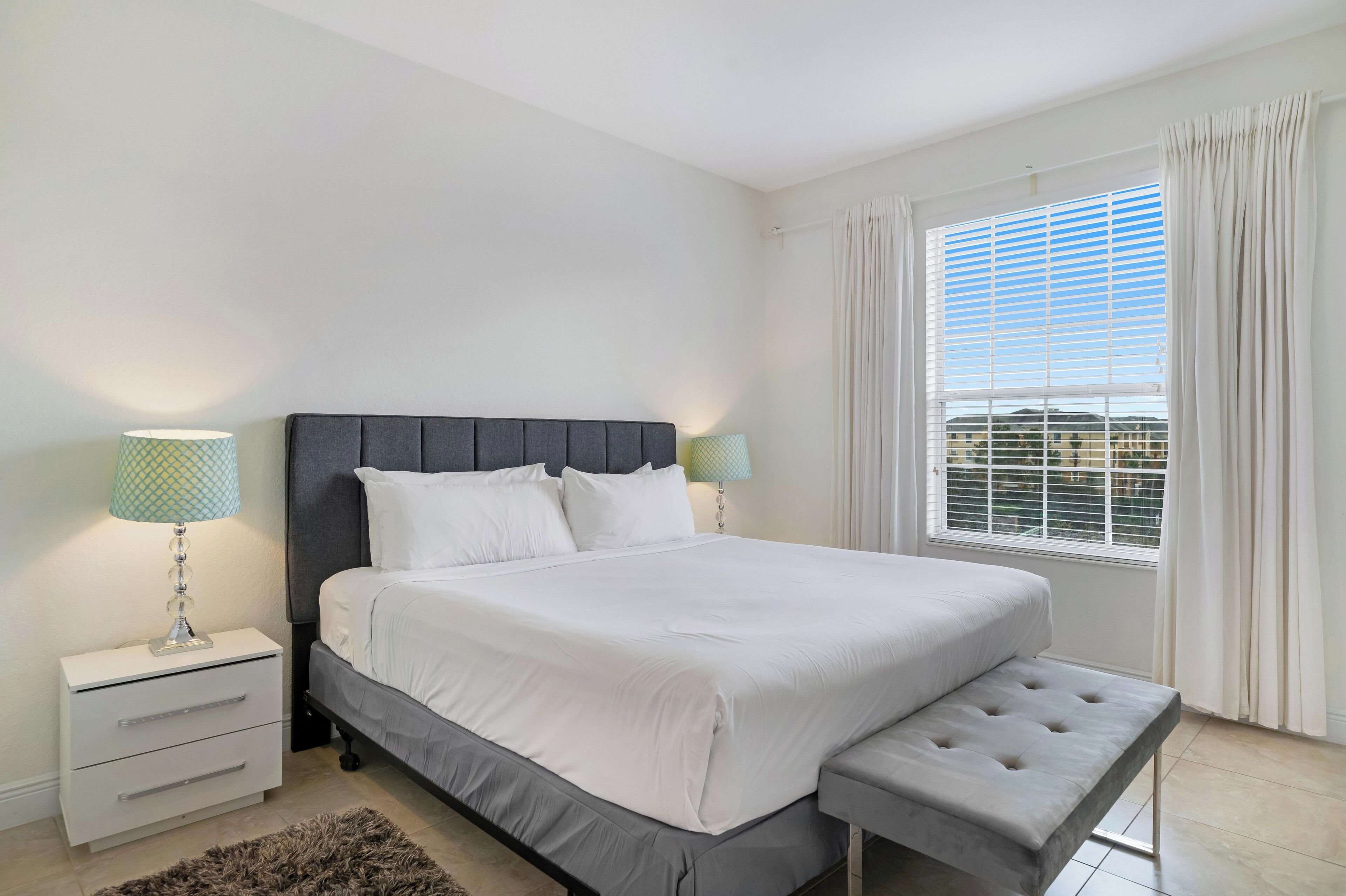 A neatly arranged bedroom with a large bed, bedside lamps, a bench, and a window with curtains.