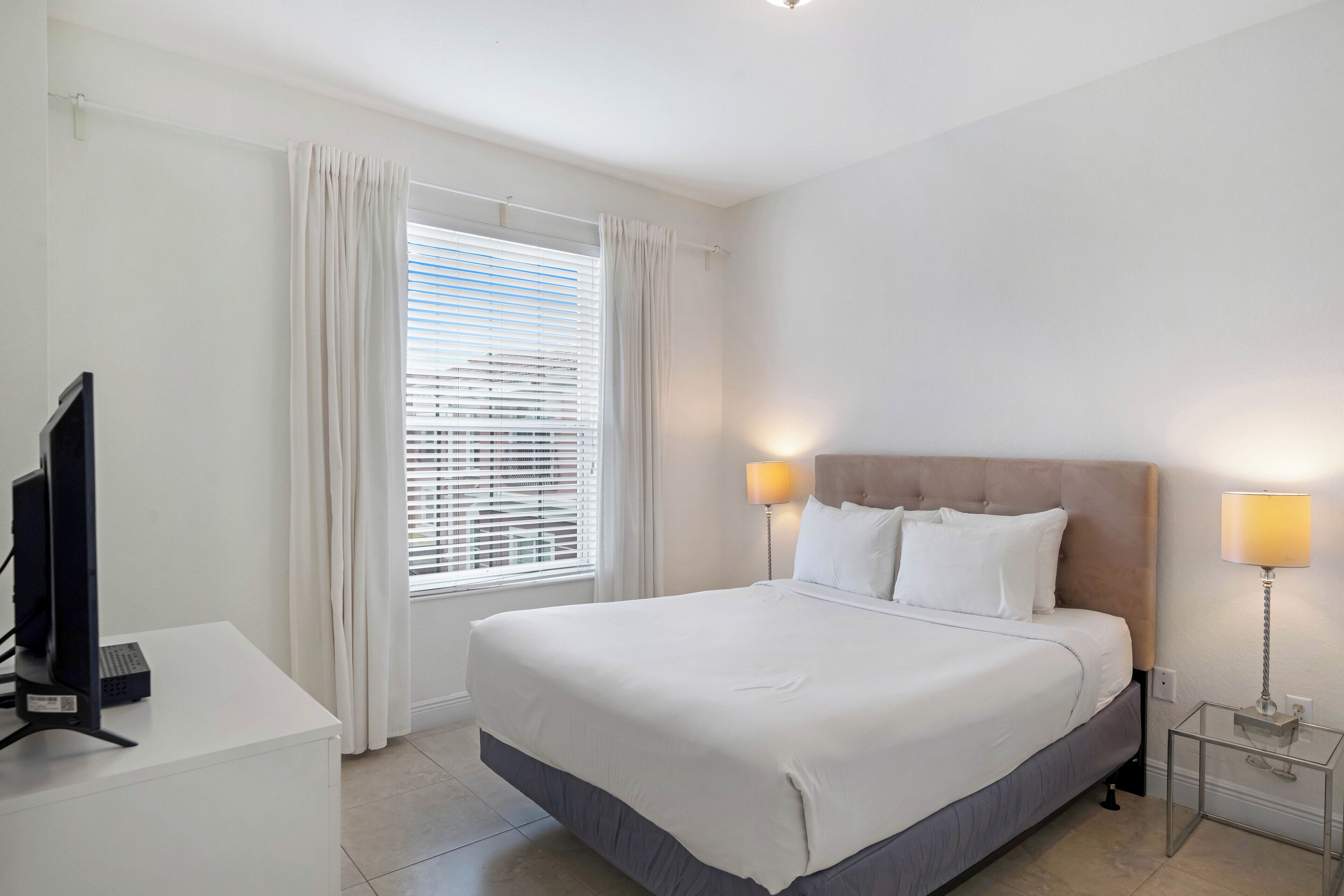A hotel room with a large bed, a desk with a computer, and a window with blinds.