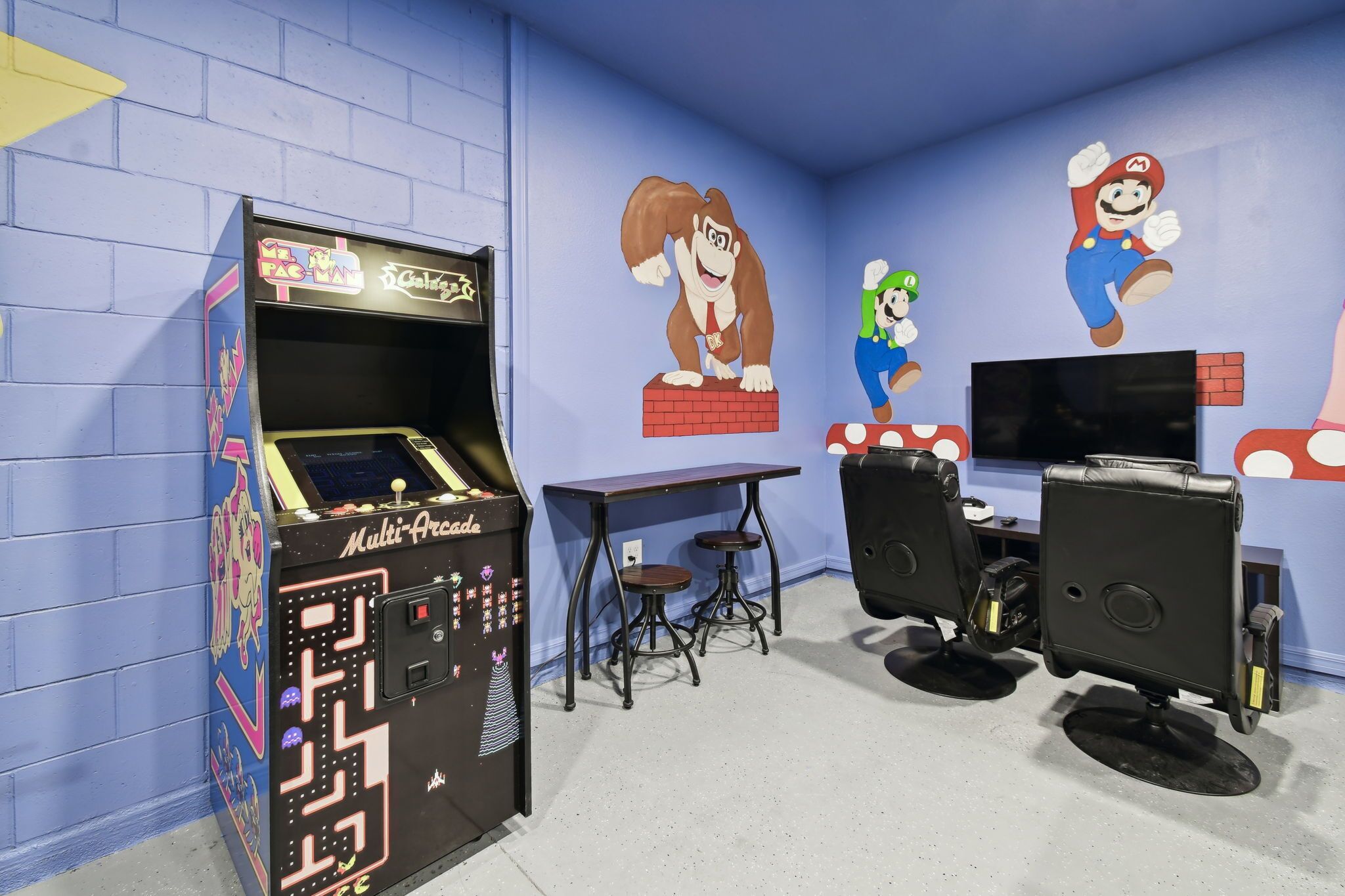 Ett rum med en spelmaskin, två pallar, en tv och en vägg dekorerad med Mario-motiv.