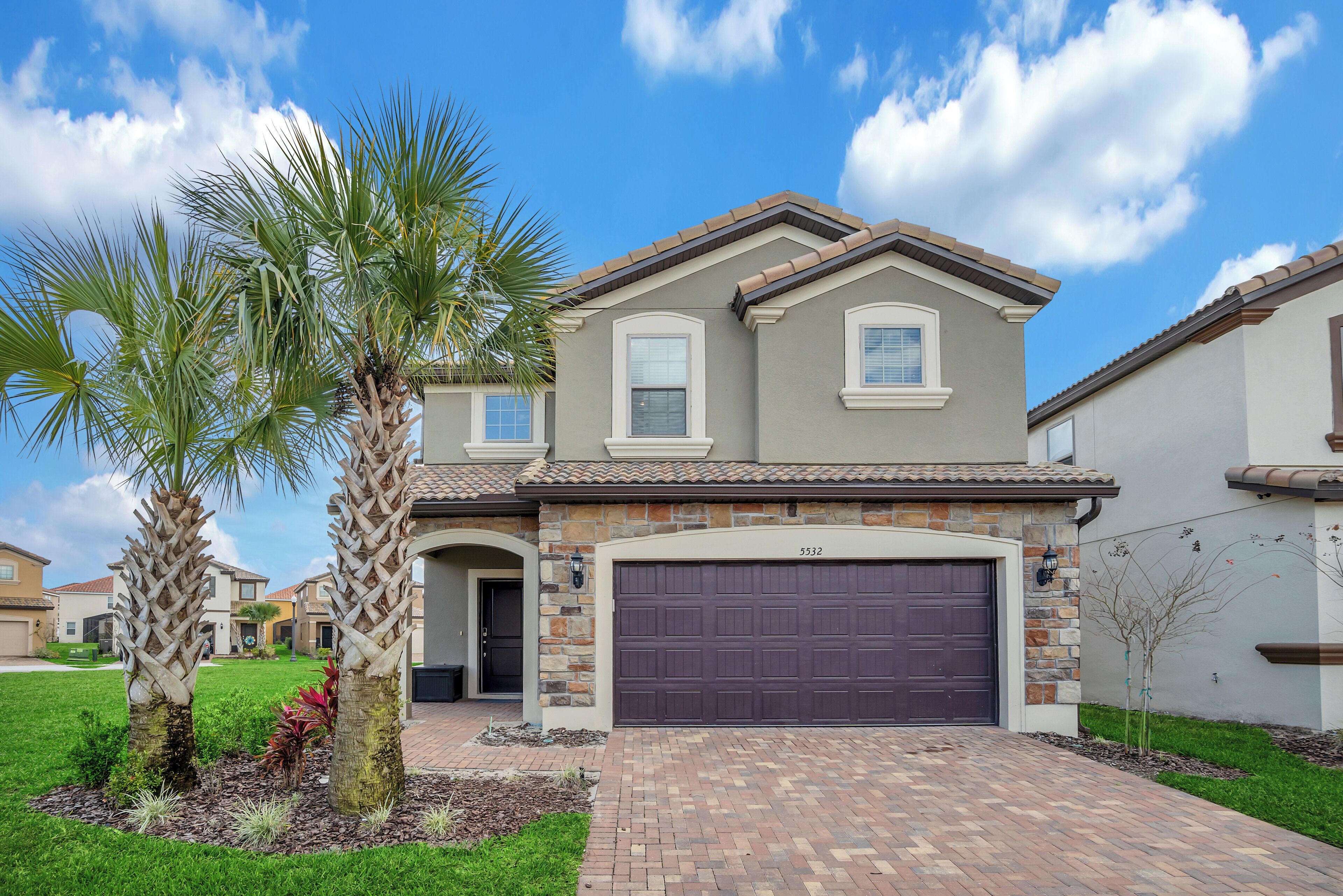 A two-story house with a stone facade, a dark garage door, and a well-maintained lawn with palm trees.