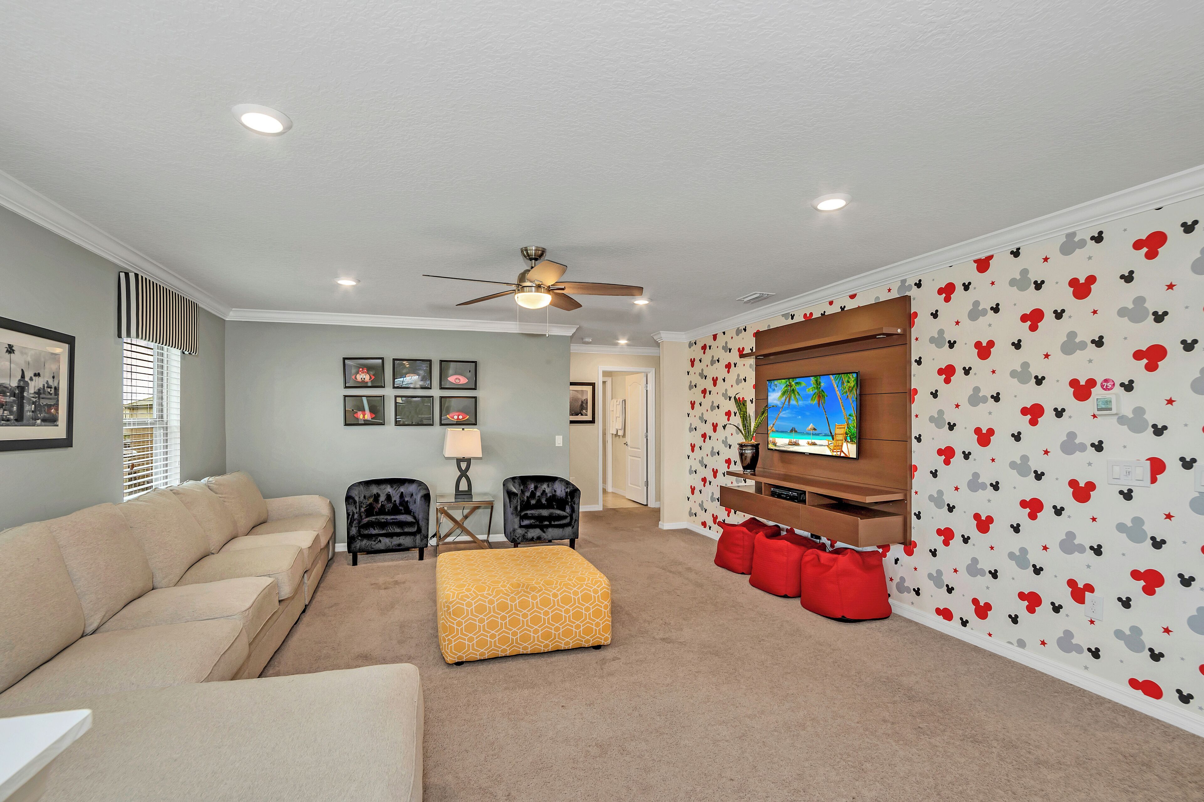 A living room with a beige sectional sofa, a yellow ottoman, a ceiling fan, a TV on a wooden stand, and Mickey Mouse themed wallpaper.