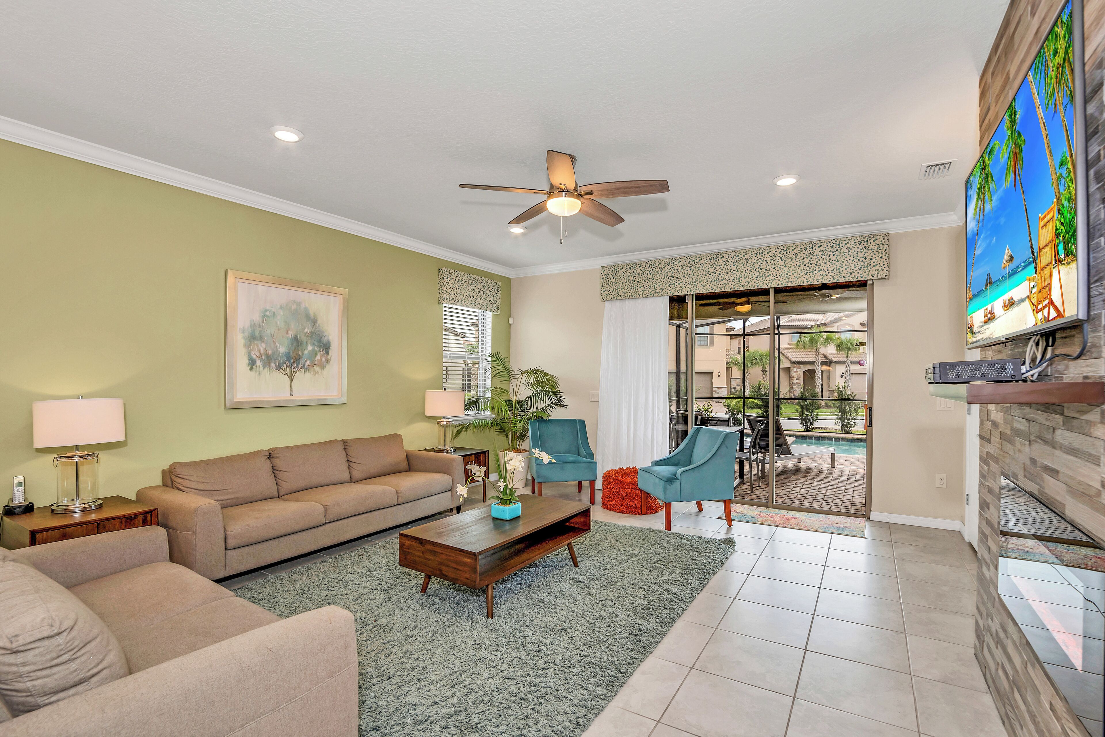 A living room with a sofa, coffee table, and chairs, a ceiling fan, and a TV on the wall.
