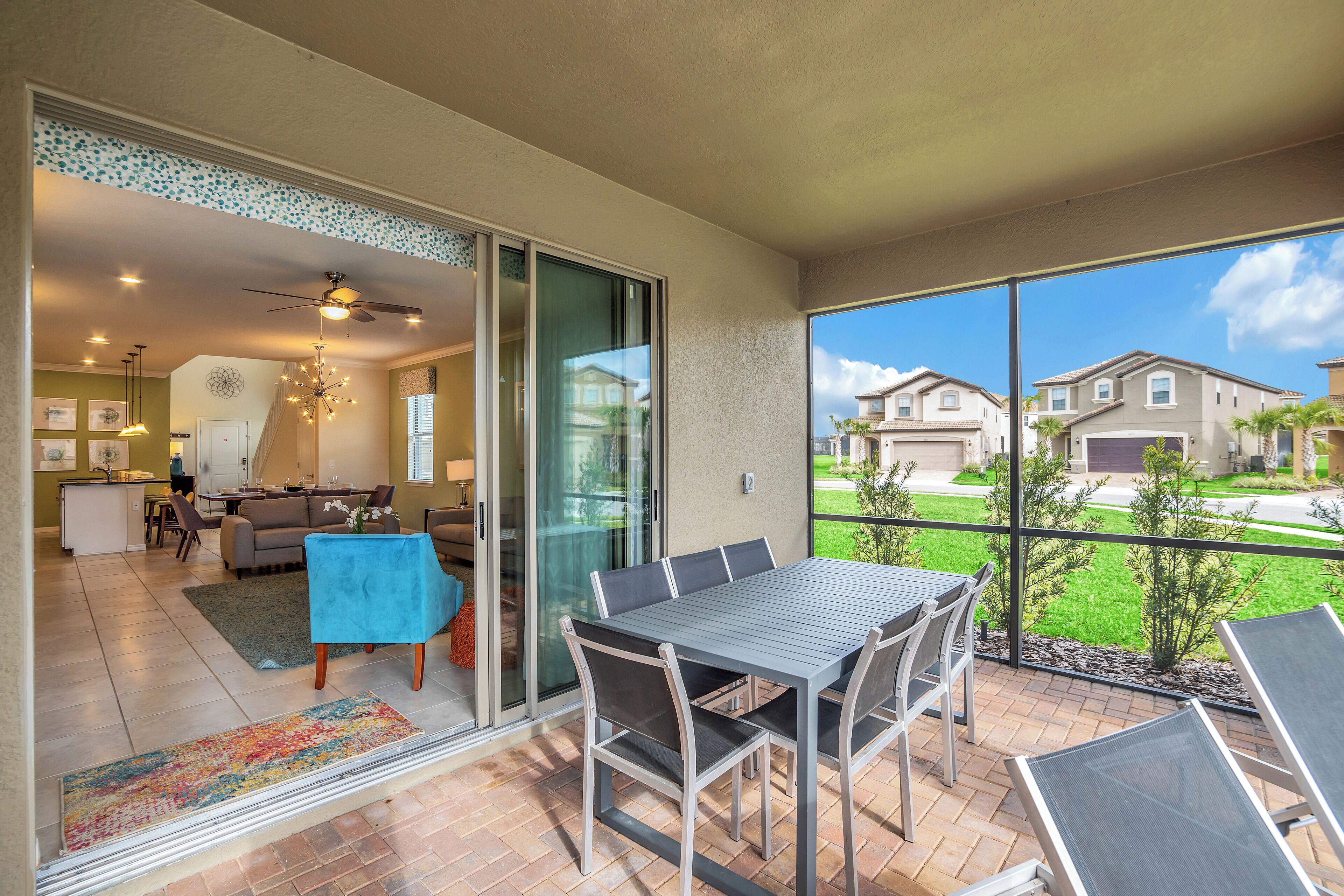 A spacious balcony with a dining table and chairs, a sliding glass door leading to a living area, and a clear view of neighboring houses.
