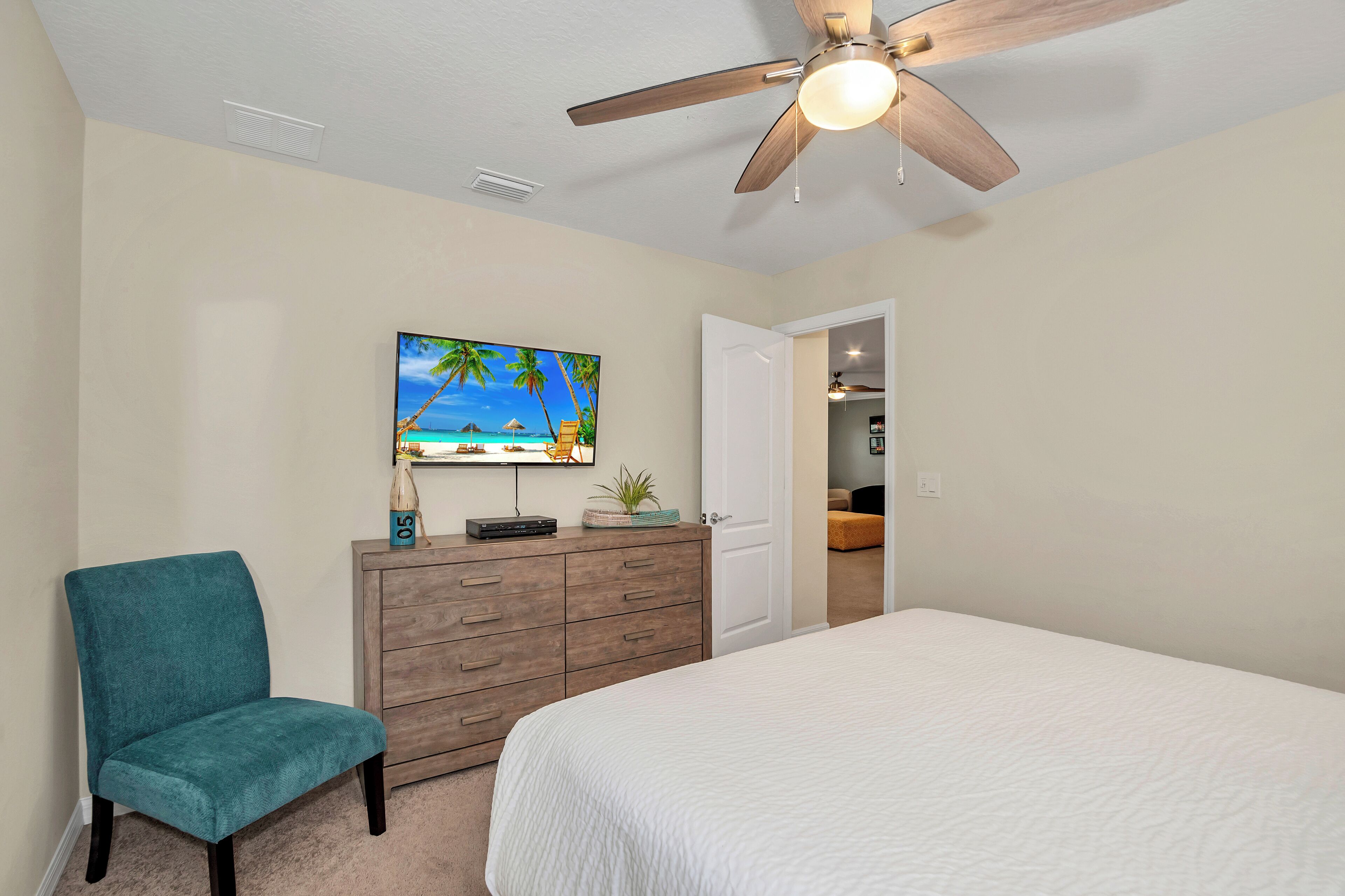 Une chambre à coucher avec un lit, un fauteuil de couleur turquoise, une commode en bois, un ventilateur de plafond et un téléviseur fixé au mur.
