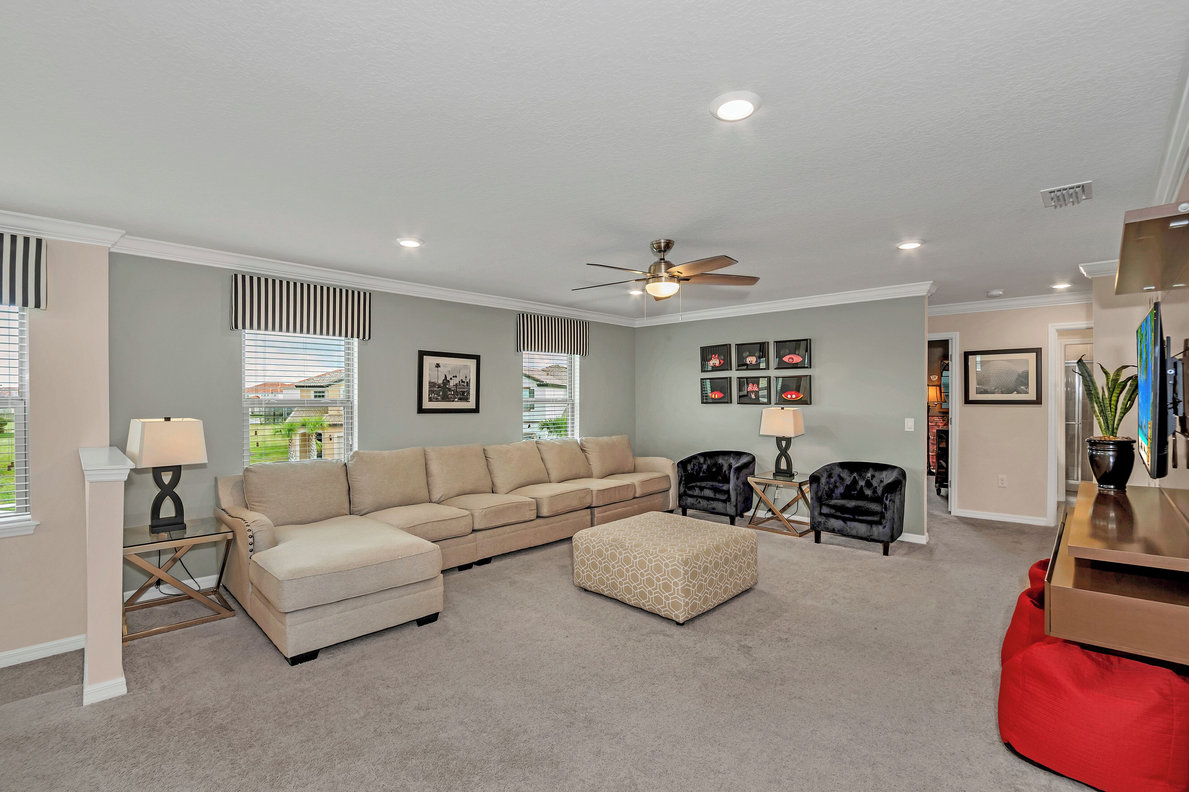 A living room with a beige sectional sofa, a matching ottoman, a black armchair, a red bean bag, and a ceiling fan.
