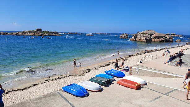 Une plage avec une eau d’un bleu limpide, des affleurements rocheux et plusieurs bateaux gonflables sur le sable.