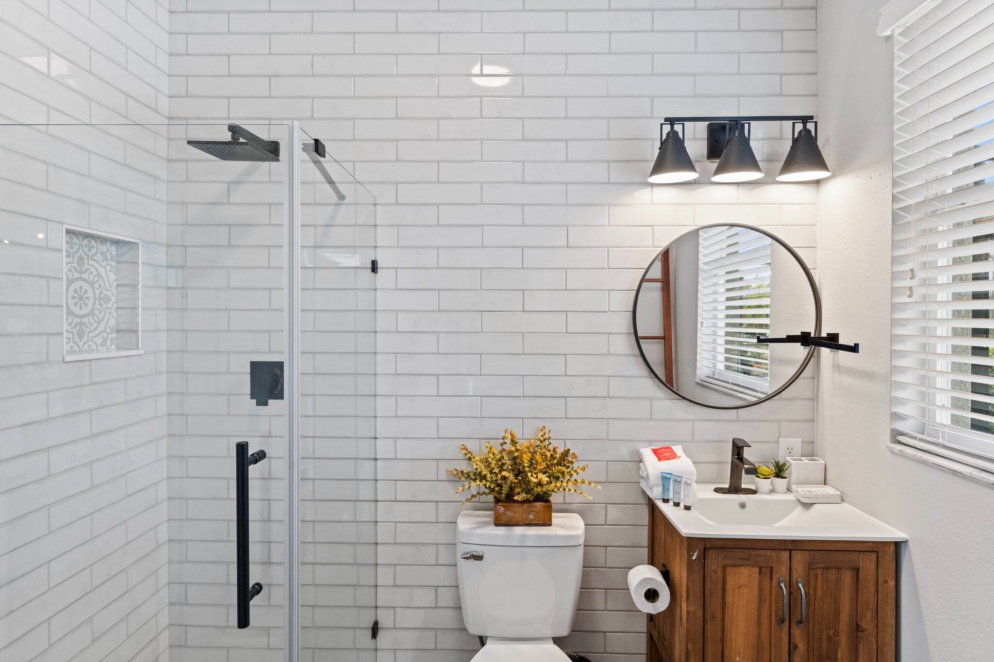 Une salle de bain avec des carreaux de céramique blancs de type « métro », une cabine de douche en verre, un meuble en bois, un miroir rond et des toilettes dotées d’un distributeur d’essuie-tout.