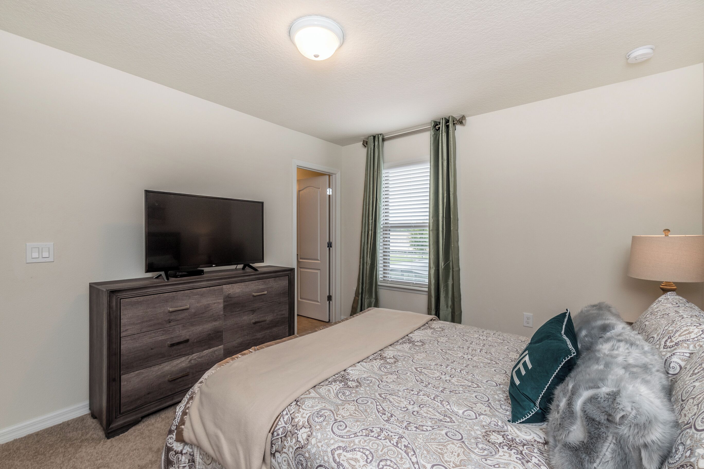 A bedroom with a bed, a dresser, a television, a window with curtains, and a lamp.