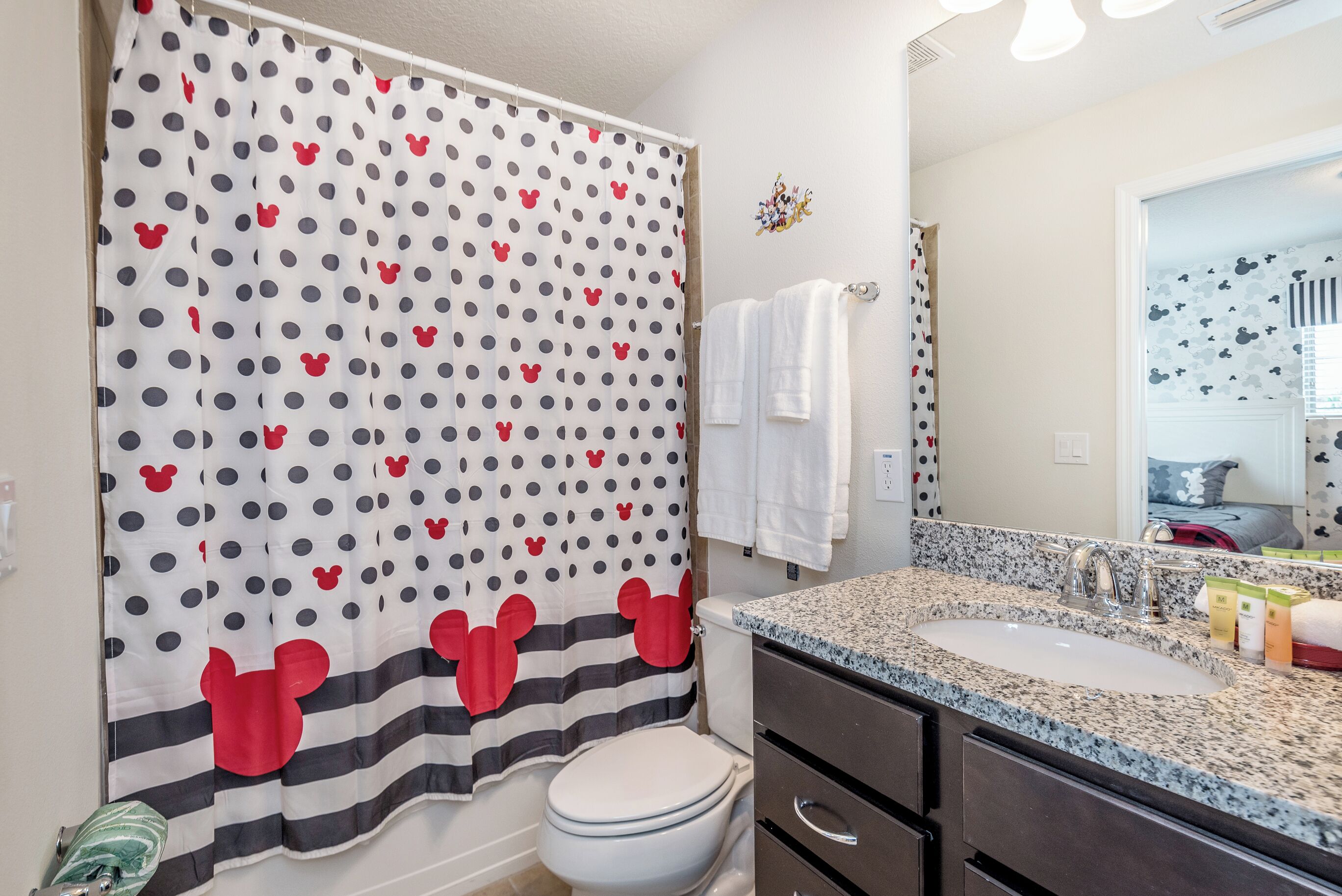 A bathroom with a Mickey Mouse themed shower curtain, a white toilet, a granite countertop sink, and a mirror reflecting the bathroom.