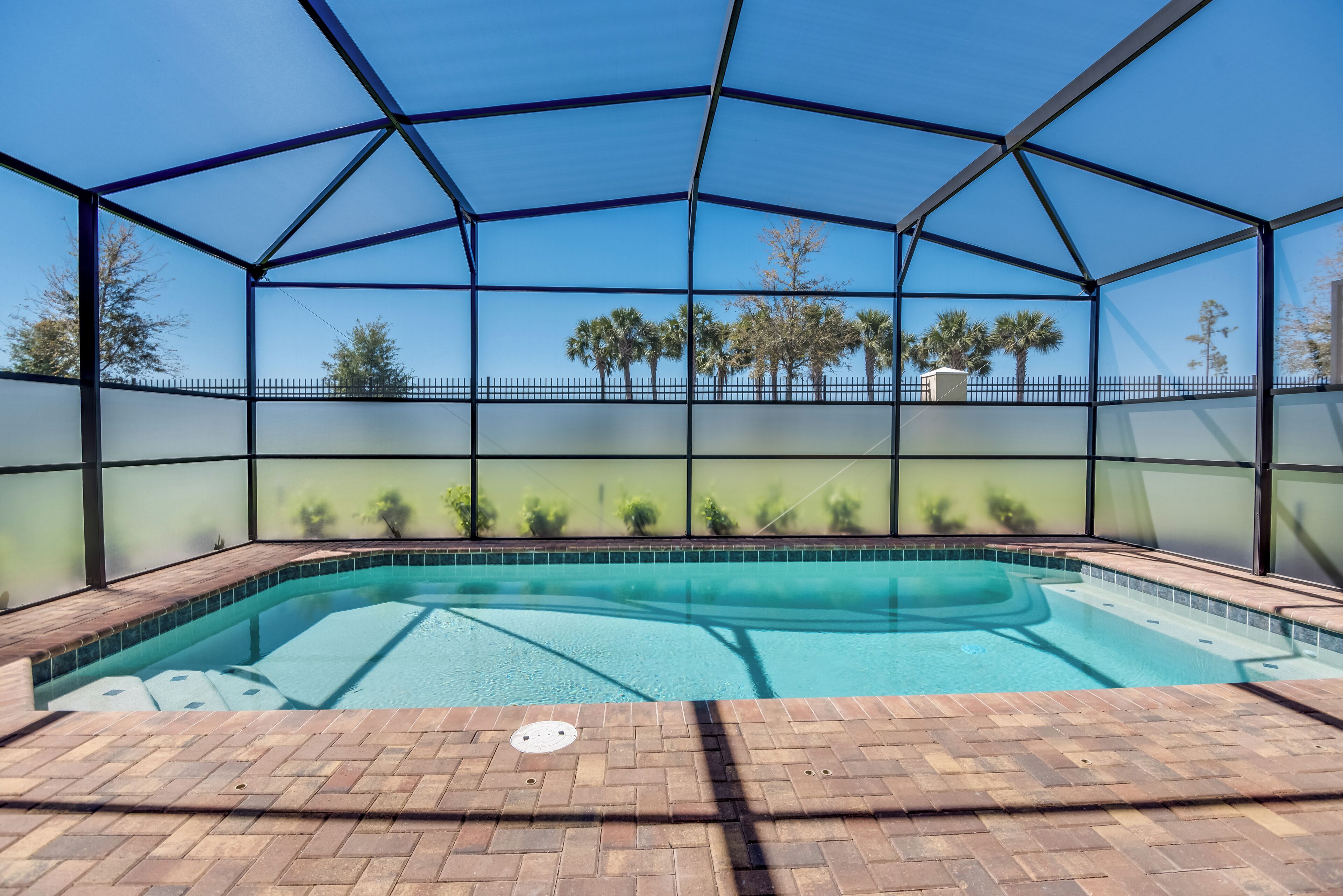 An indoor swimming pool with a clear view of palm trees and a fenced area.