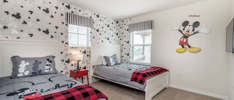 A bedroom with two beds, Mickey Mouse wall decal, and striped window valances.