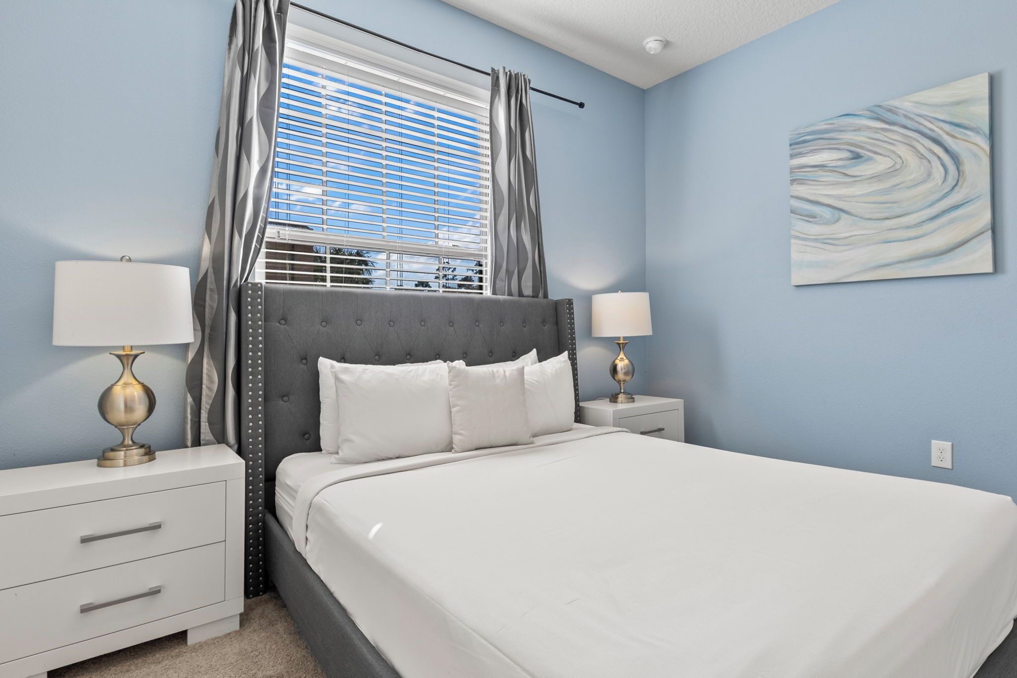 A bedroom with a bed, two bedside tables, a window with curtains, and a wall-mounted artwork.