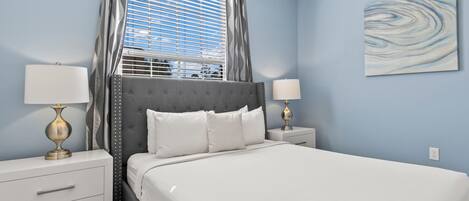 A bedroom with a bed, two bedside tables, a window with curtains, and a wall-mounted artwork.