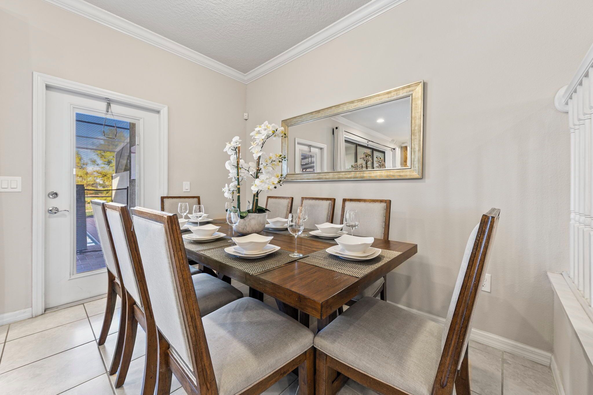 Un comedor con una mesa de madera dispuesta para seis personas, flanqueada por sillas con asientos acolchados. Un espejo grande cuelga sobre la mesa, y un jarrón con flores blancas está colocado en la mesa.