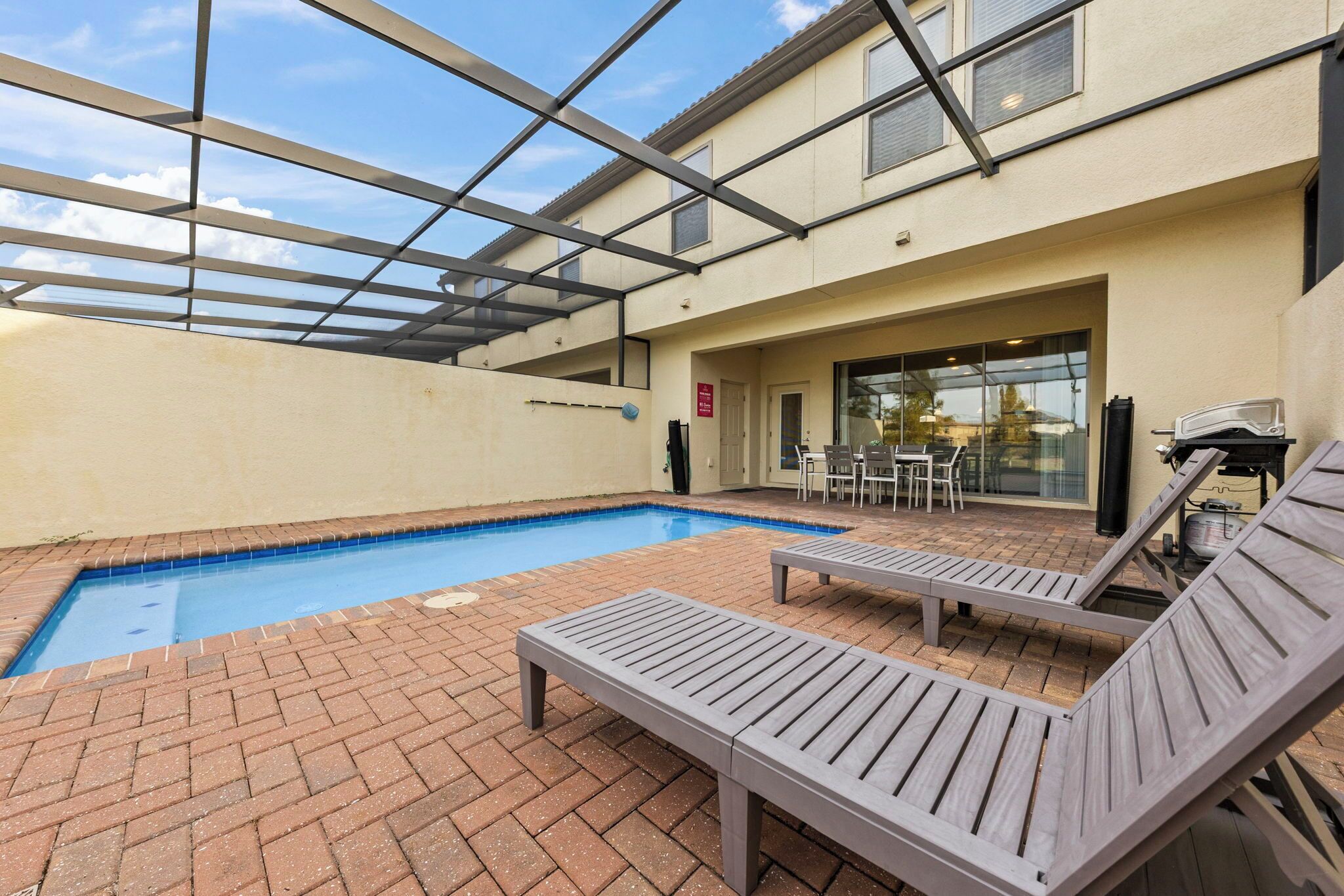Uma área de piscina com cobertura de vidro, espreguiçadeiras e um alpendre com mesa e cadeiras.