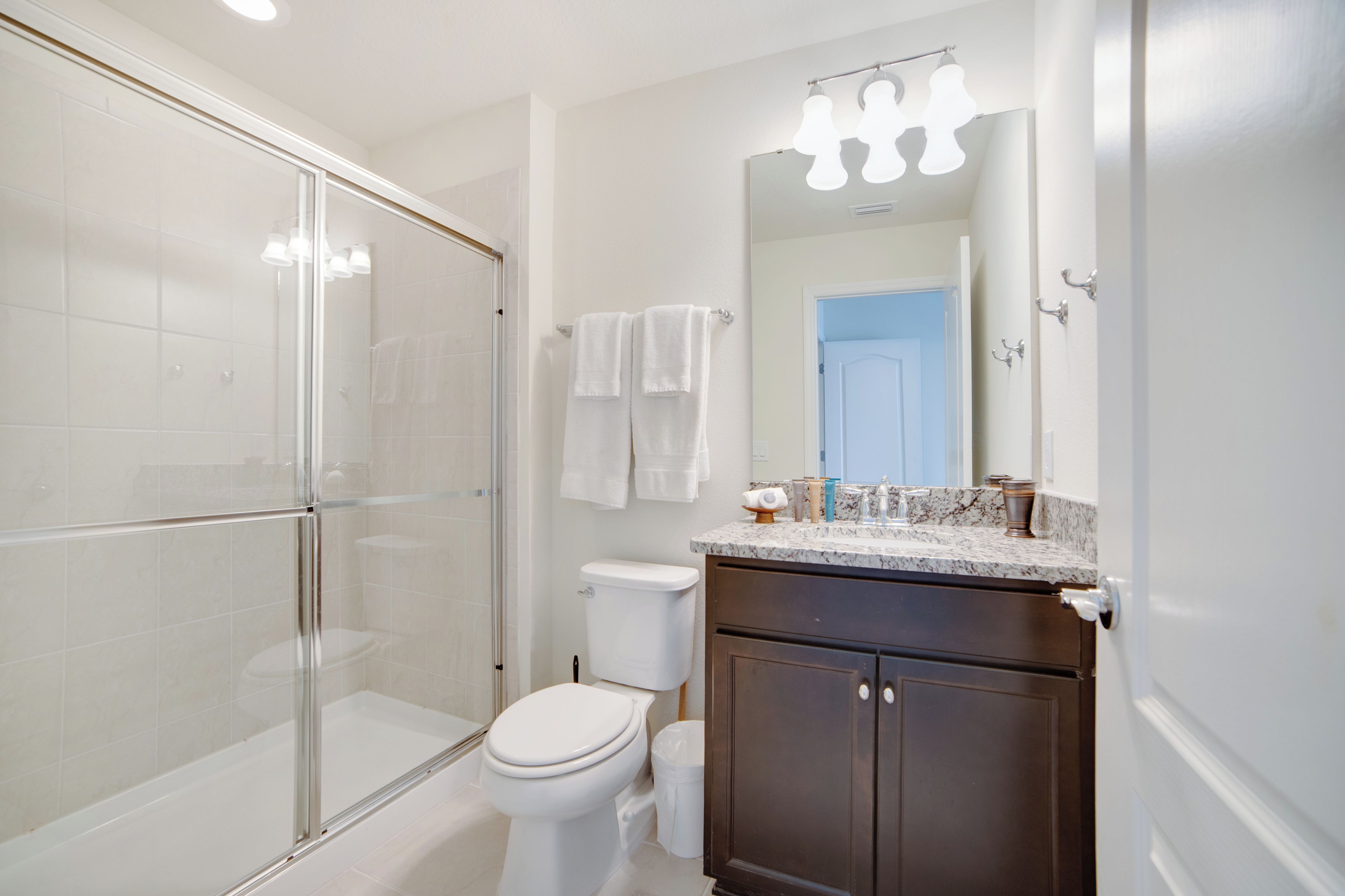 Une salle de bain moderne avec une douche à parois de verre, des toilettes blanches, un meuble-lavabo en bois foncé et un miroir avec des lumières au-dessus.