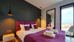 Ein modernes Schlafzimmer mit einem großen Bett, violetter Bettwäsche und Blick auf die Stadt durch ein großes Fenster.