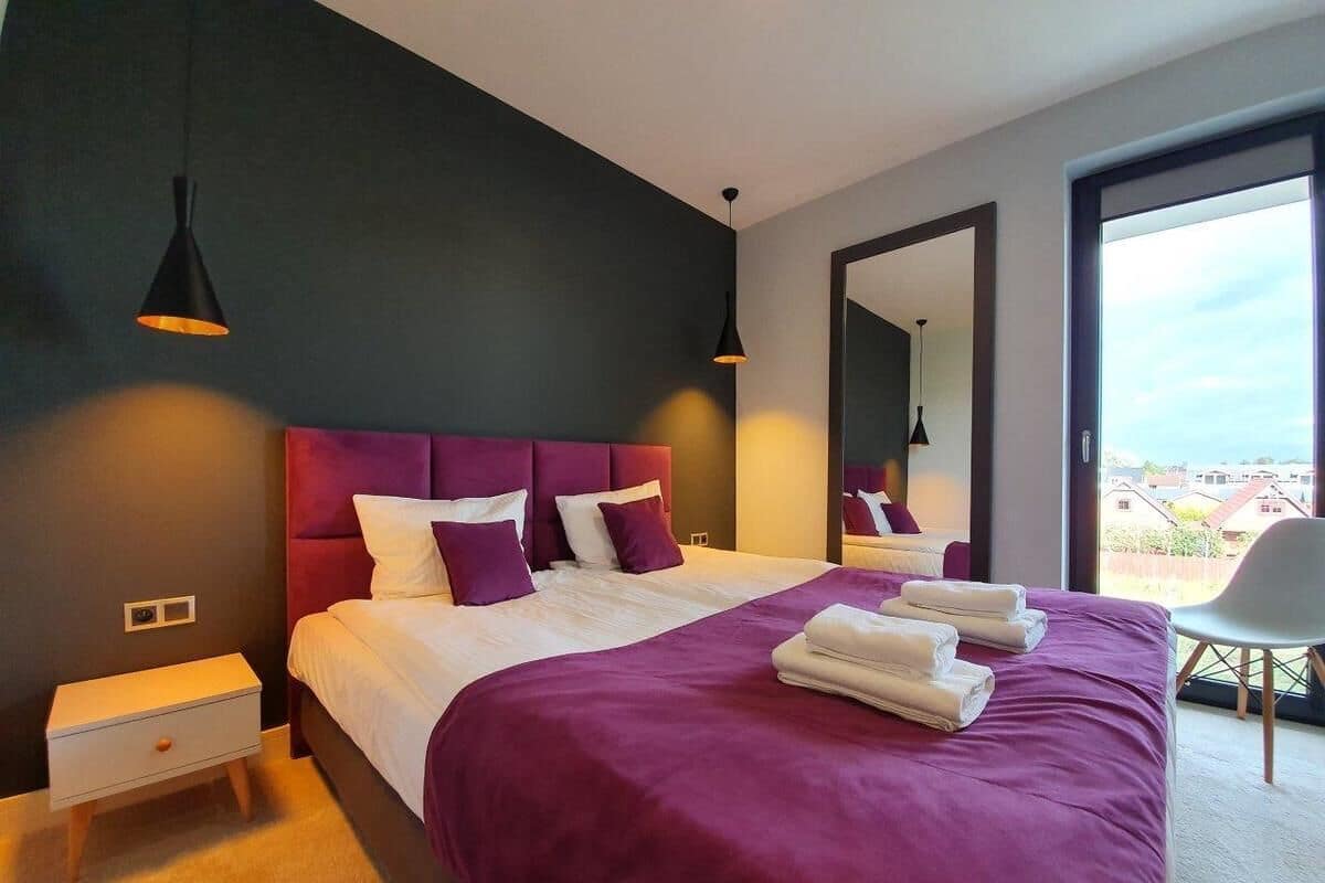 A modern bedroom with a large bed, purple bedding, and a view of the city through a large window.