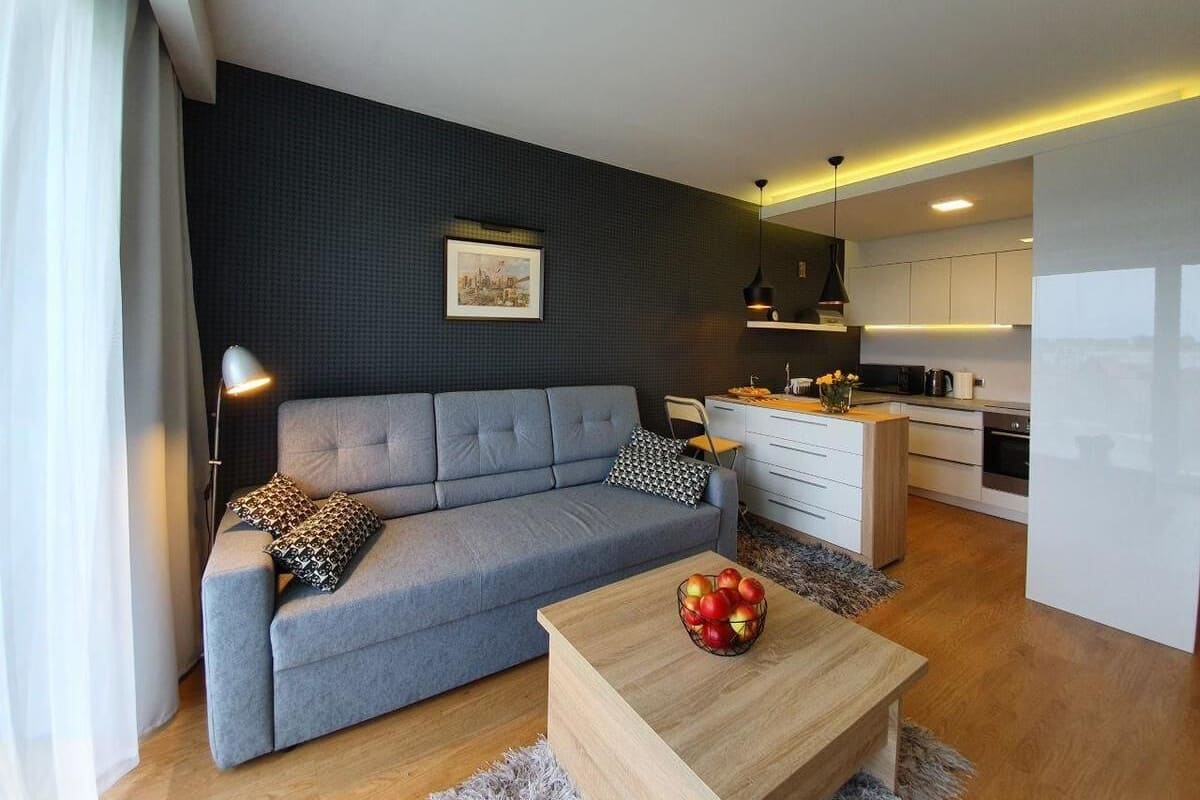 A modern living room with a grey sofa, a wooden coffee table with apples, and a kitchen in the background.