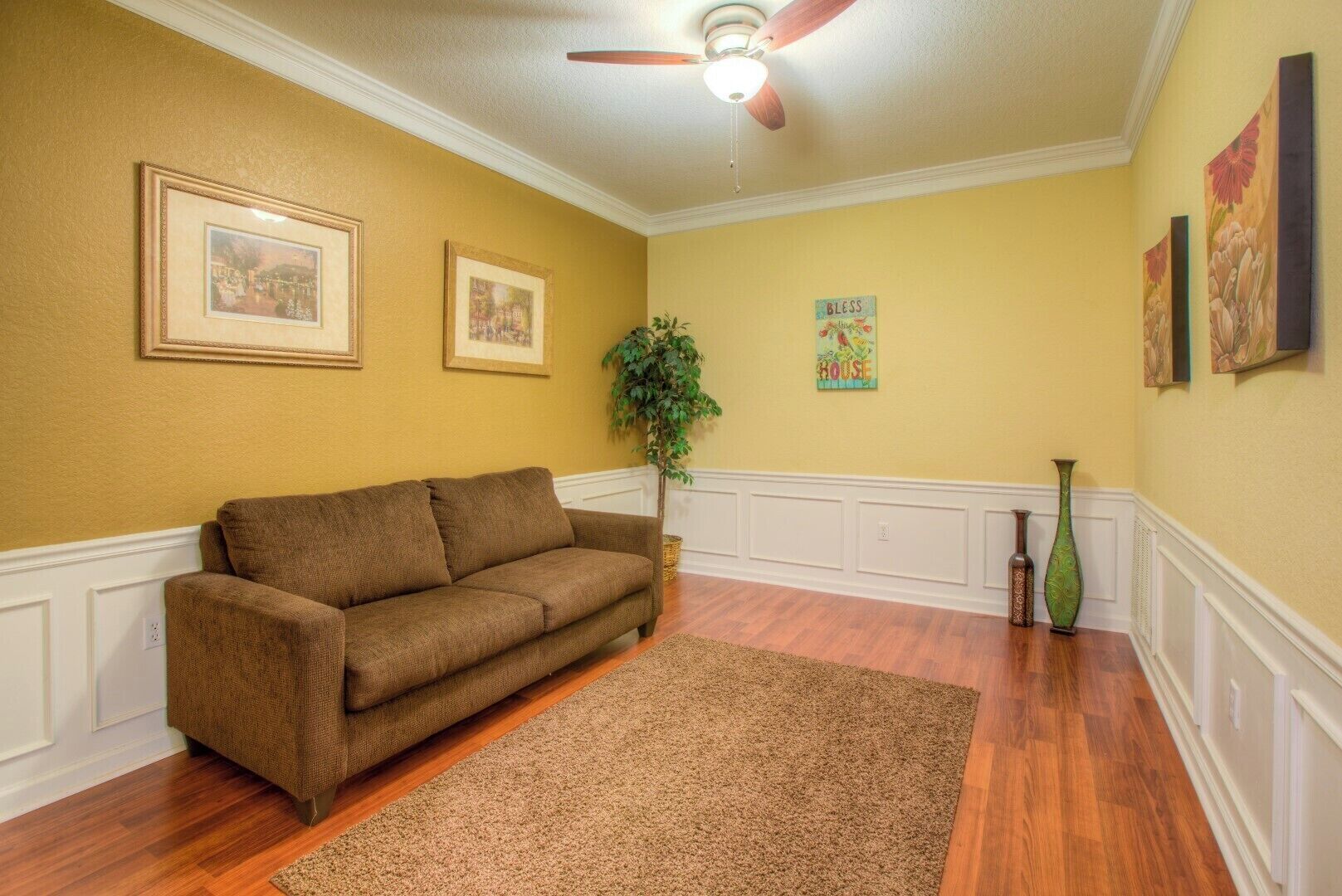 A living room with a brown sofa, wooden flooring, a ceiling fan, and framed artwork on the walls.