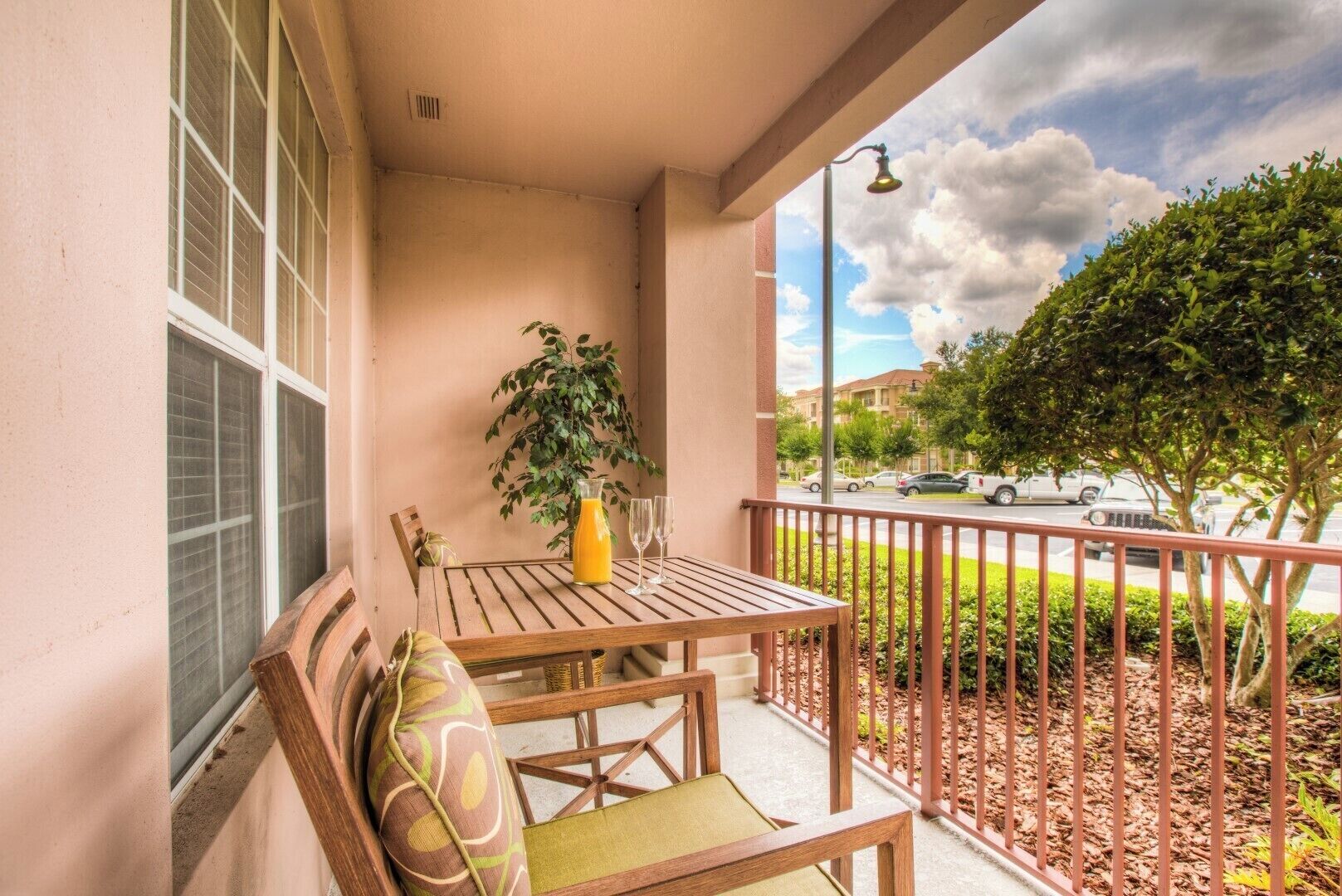 A balcony with a wooden table, two chairs, a potted plant, and a view of a street with cars and trees.