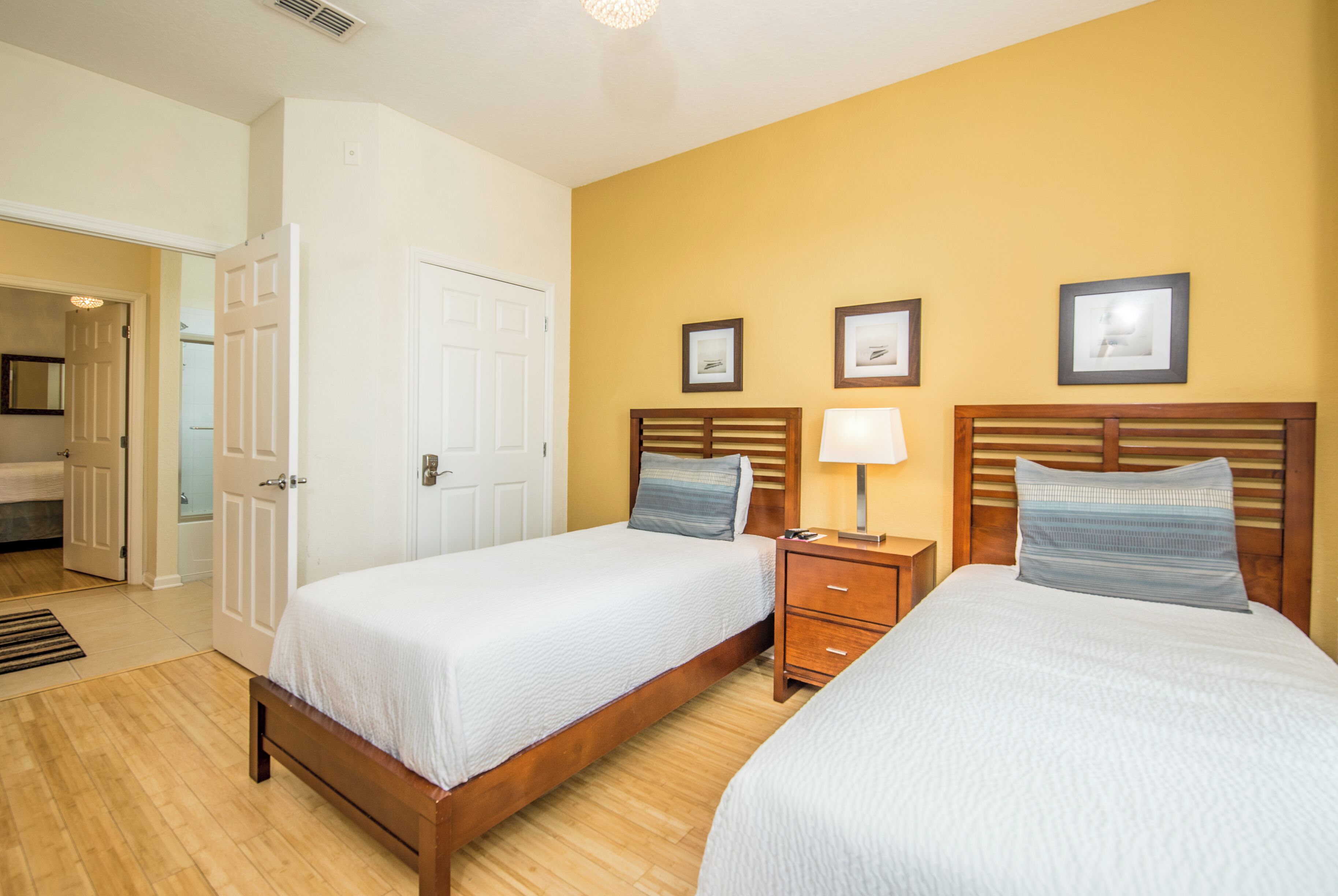 A hotel room with two beds, wooden headboards, a nightstand with a lamp, and a door leading to a bathroom.
