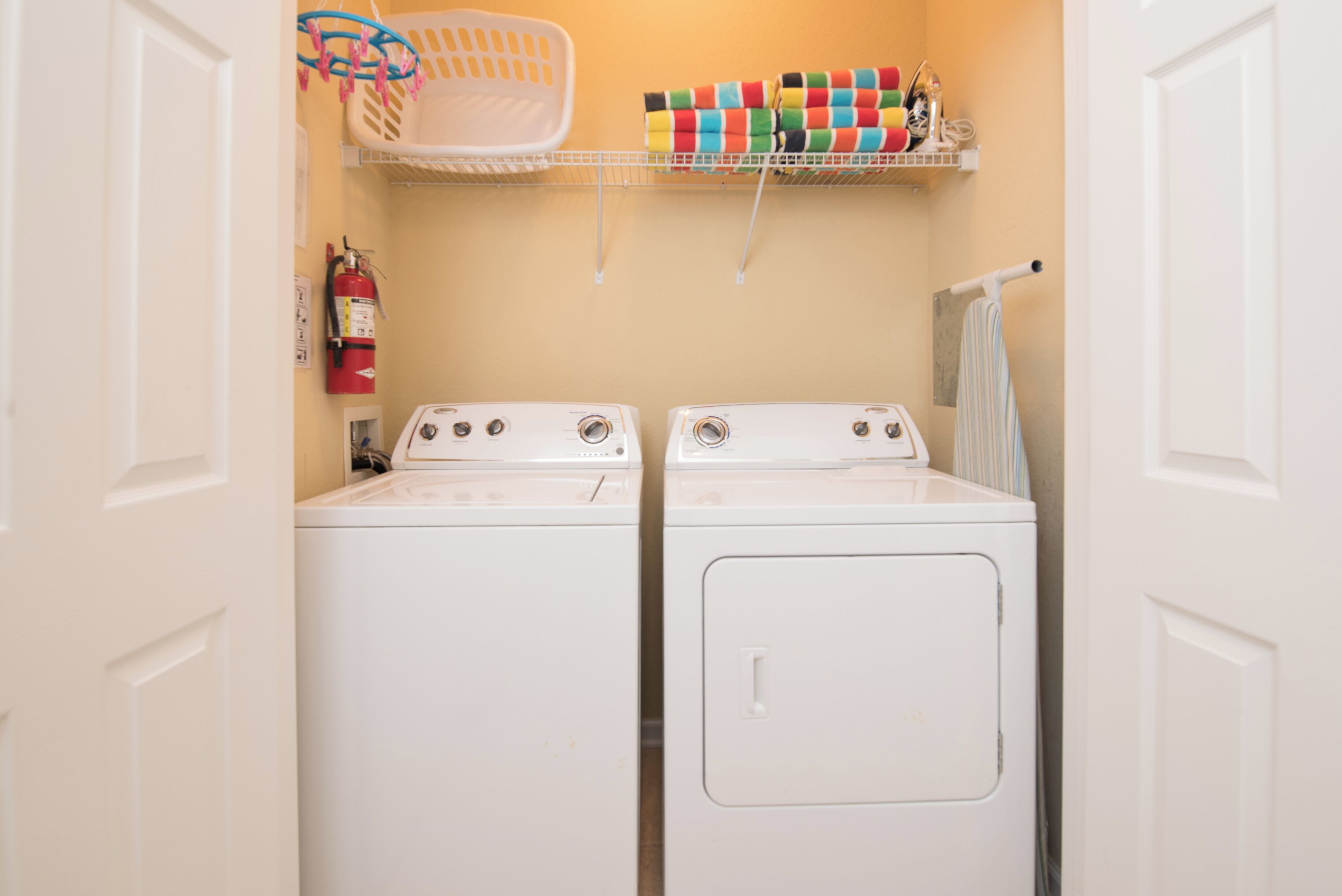 A laundry room with two white front-loading washing machines, a fire extinguisher, a laundry basket, and a shelf with colorful towels.