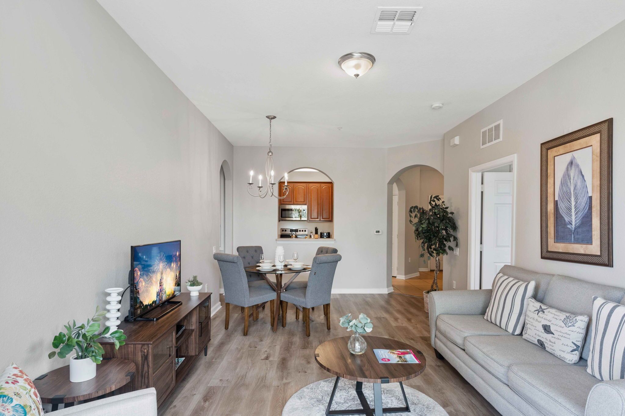 A modern living room with a grey sofa, a dining table with chairs, a TV, and a kitchen in the background.