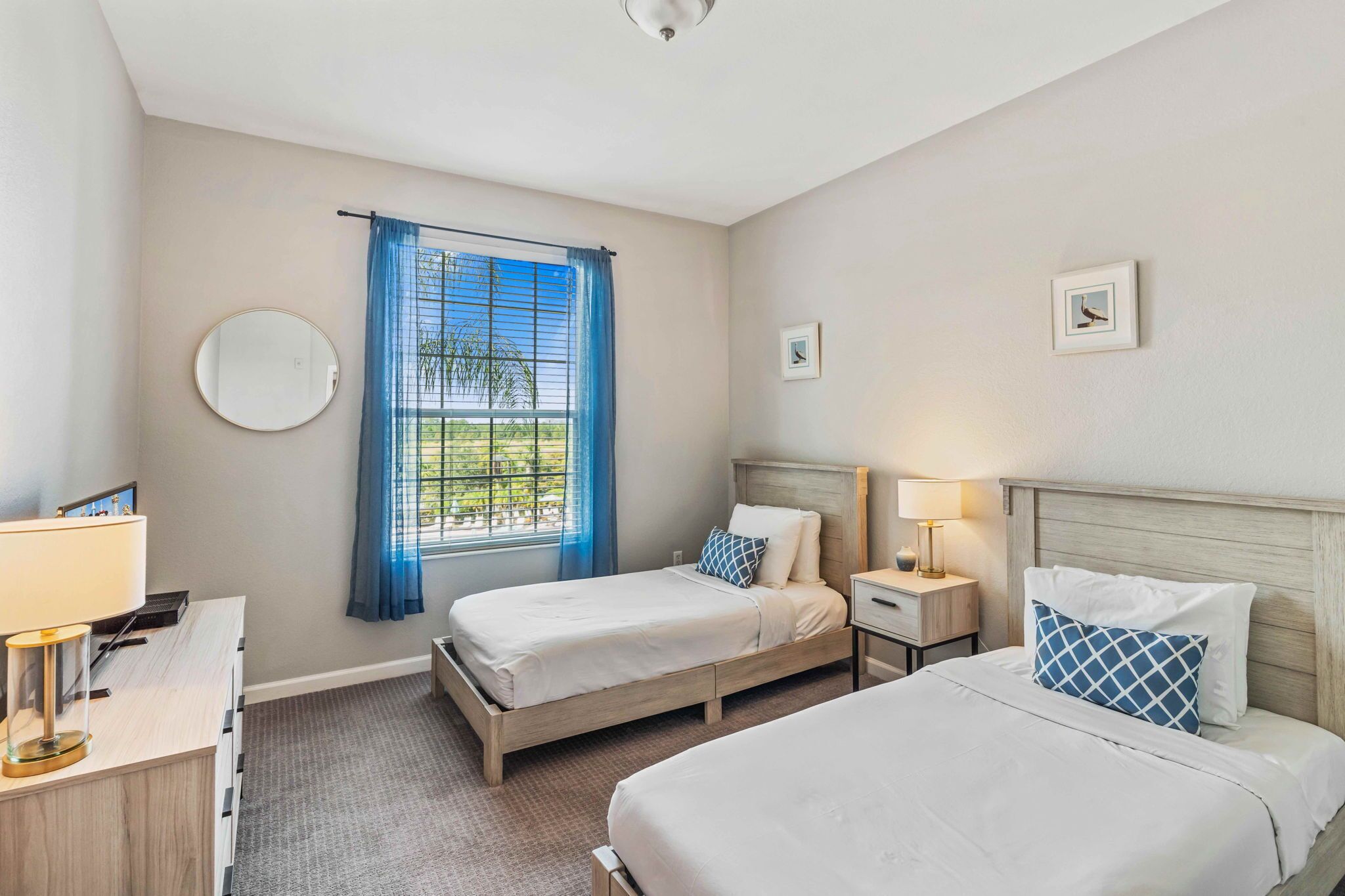 A hotel room with two beds, a round mirror, a window with blue curtains, a bedside table with a lamp, and a small picture on the wall.