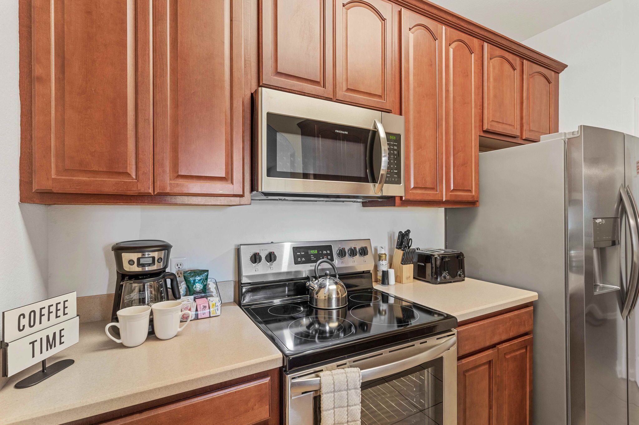 A kitchen with wooden cabinets, a stainless steel microwave, a stove, a coffee maker, and a sign that reads "COFFEE TIME".