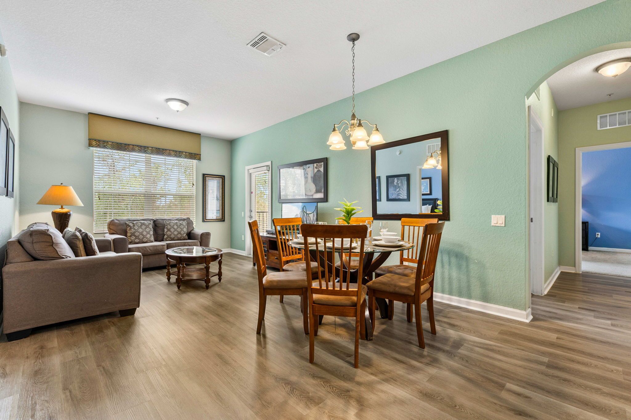 A spacious living room with a sofa, dining table, and chairs, a large mirror, and a ceiling light fixture.