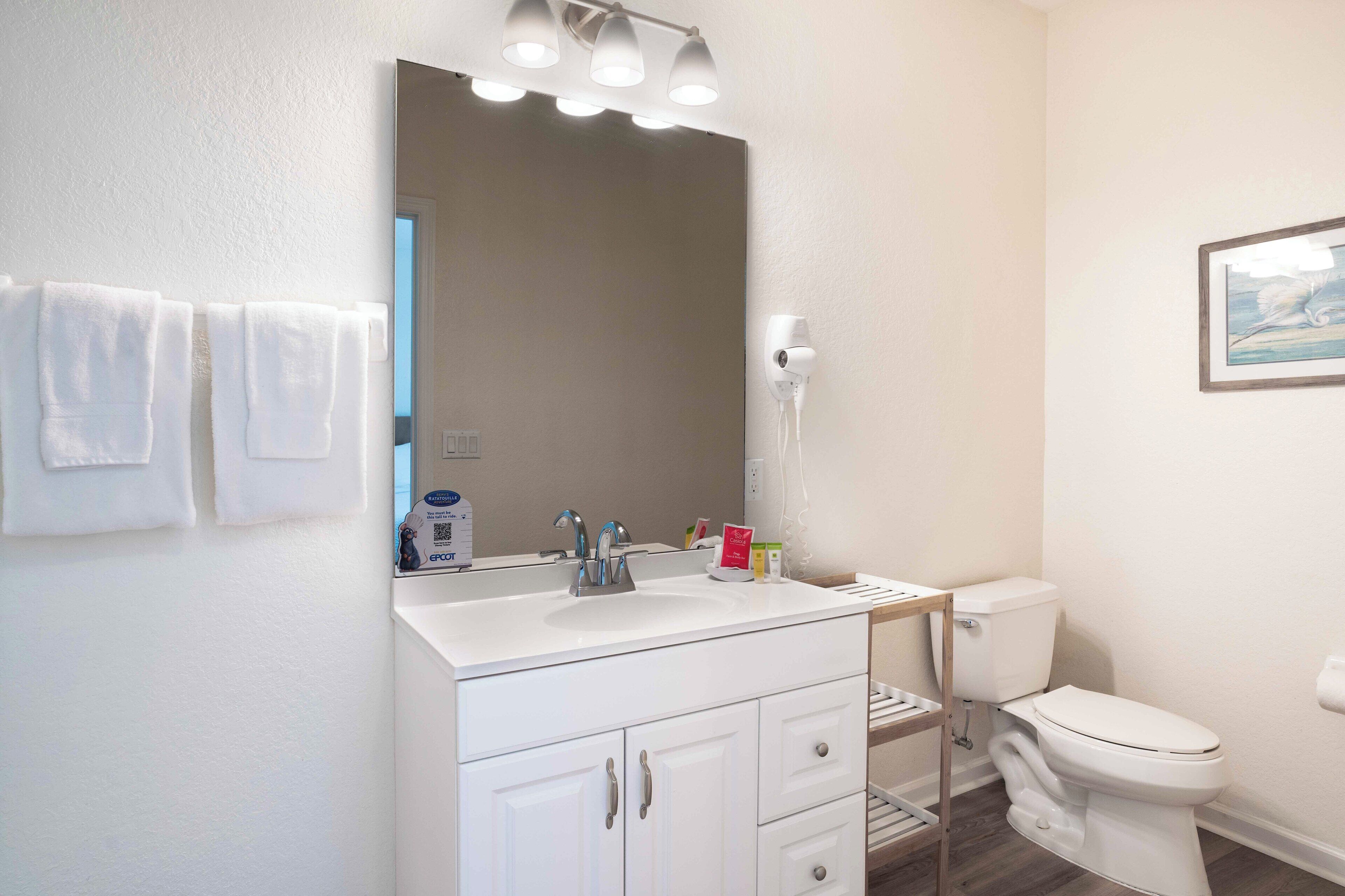 A bathroom with a white sink, mirror, and toilet. There are towels hanging on the wall and a framed picture on the wall.