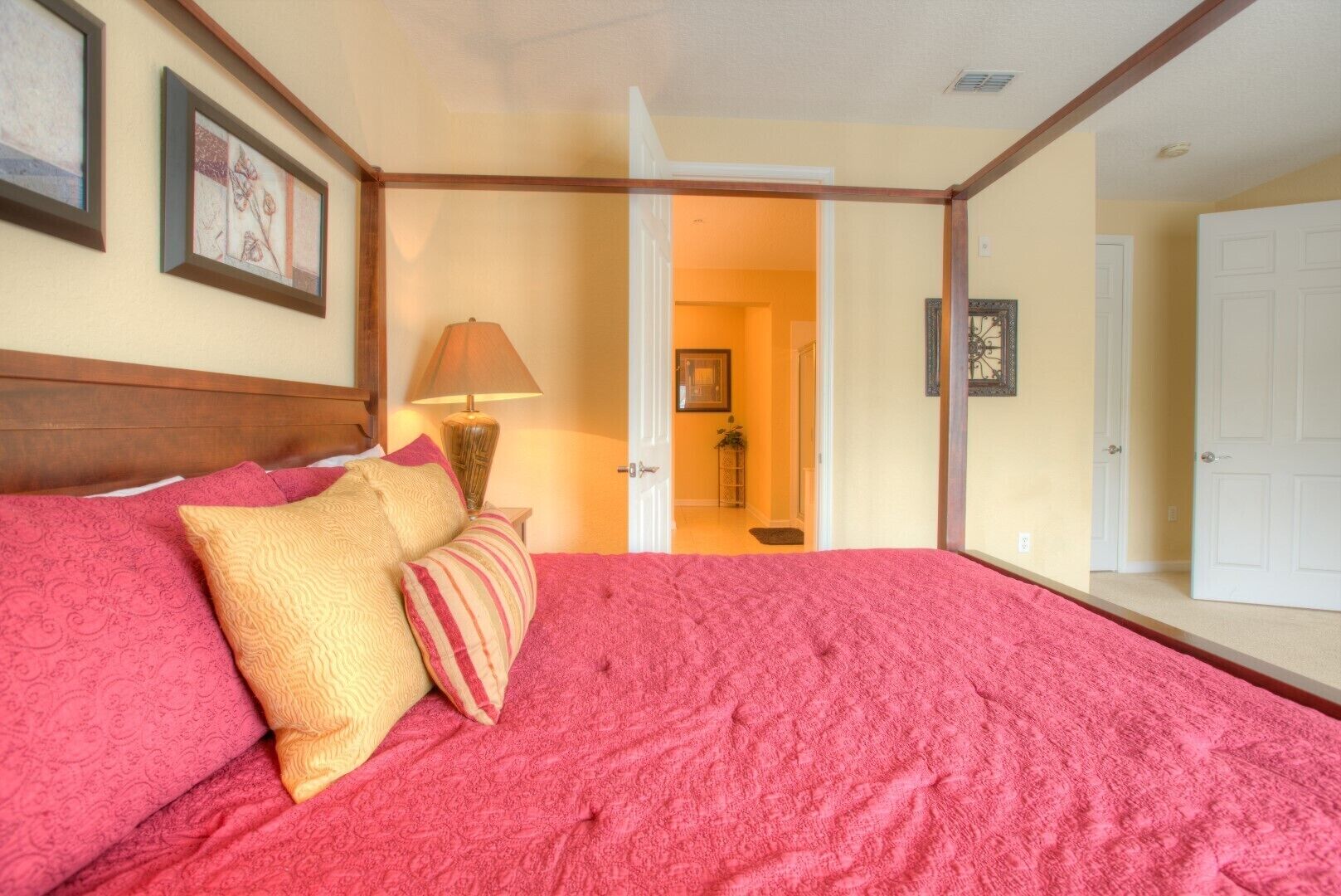 A bedroom with a large bed, a nightstand with a lamp, and a door leading to another room.