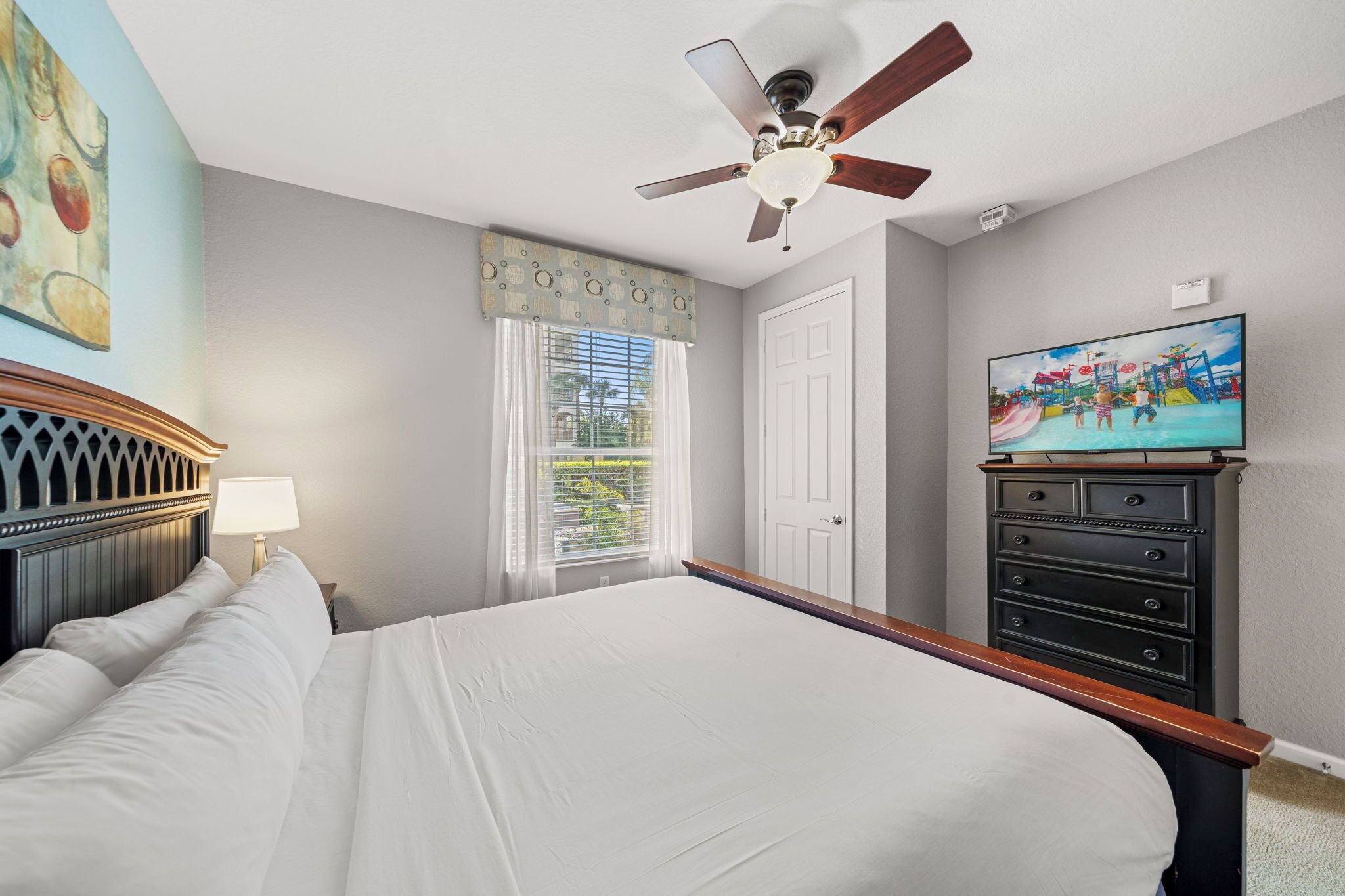 Une chambre à coucher comprenant un lit, un ventilateur de plafond, une commode, un téléviseur et une fenêtre avec des rideaux.