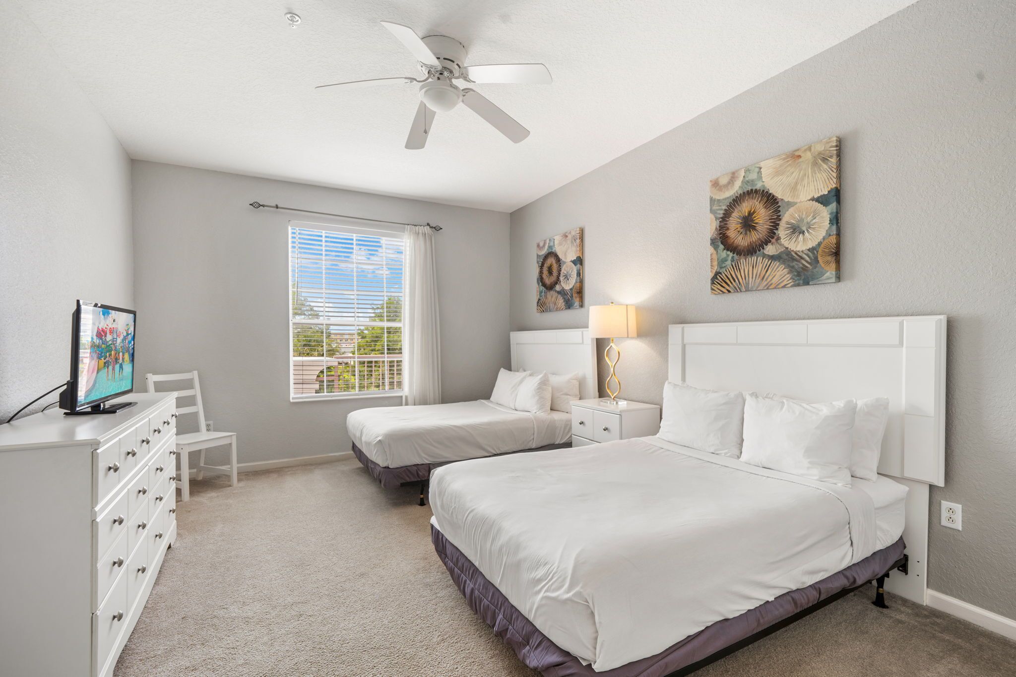 A bedroom with two beds, a ceiling fan, artwork on the walls, and a window with blinds.