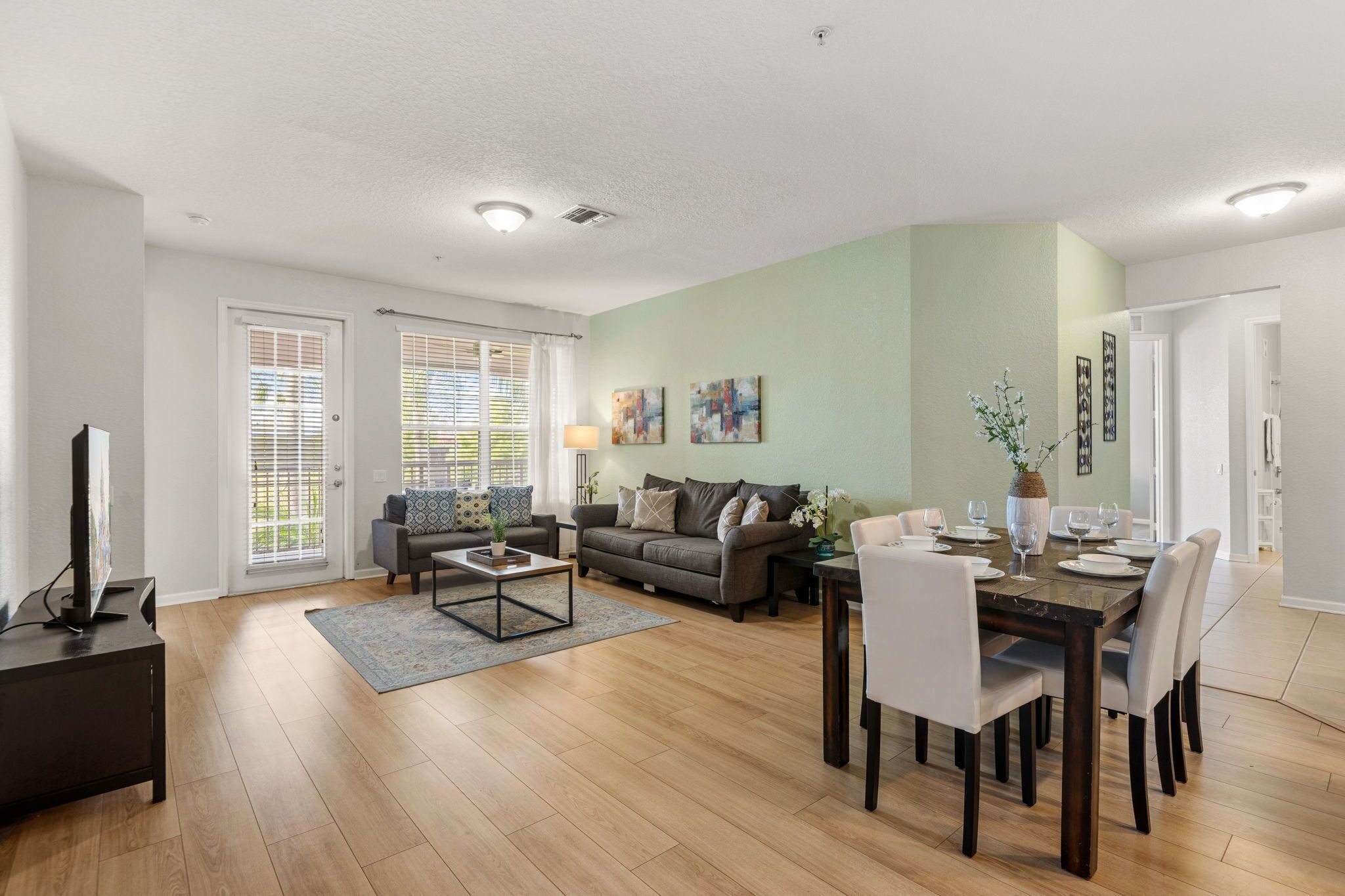 A modern living room with a dining area, hardwood flooring, and a large window with blinds.