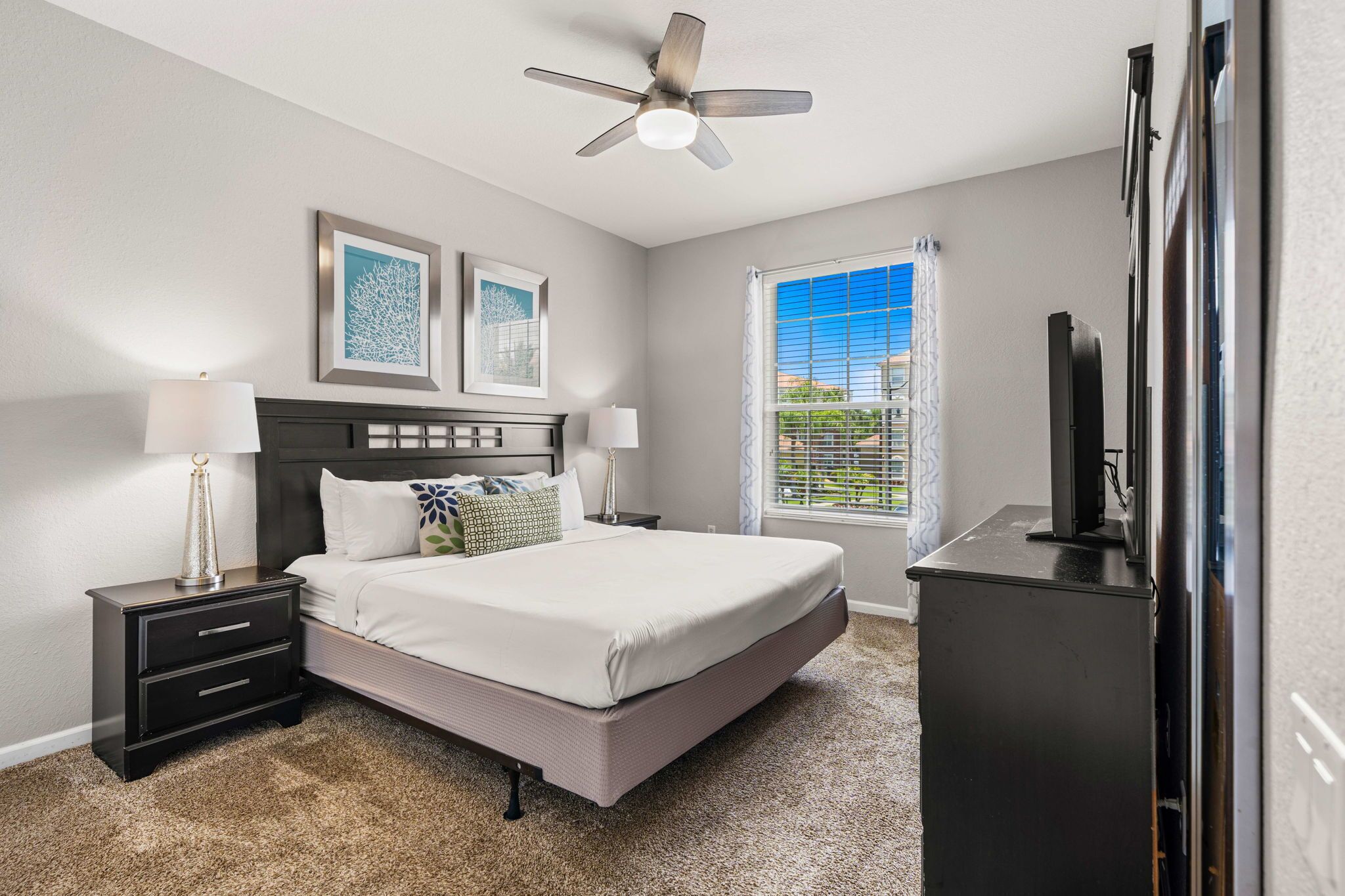 A bedroom with a bed, nightstands, a ceiling fan, artwork on the wall, a window with curtains, and a television.