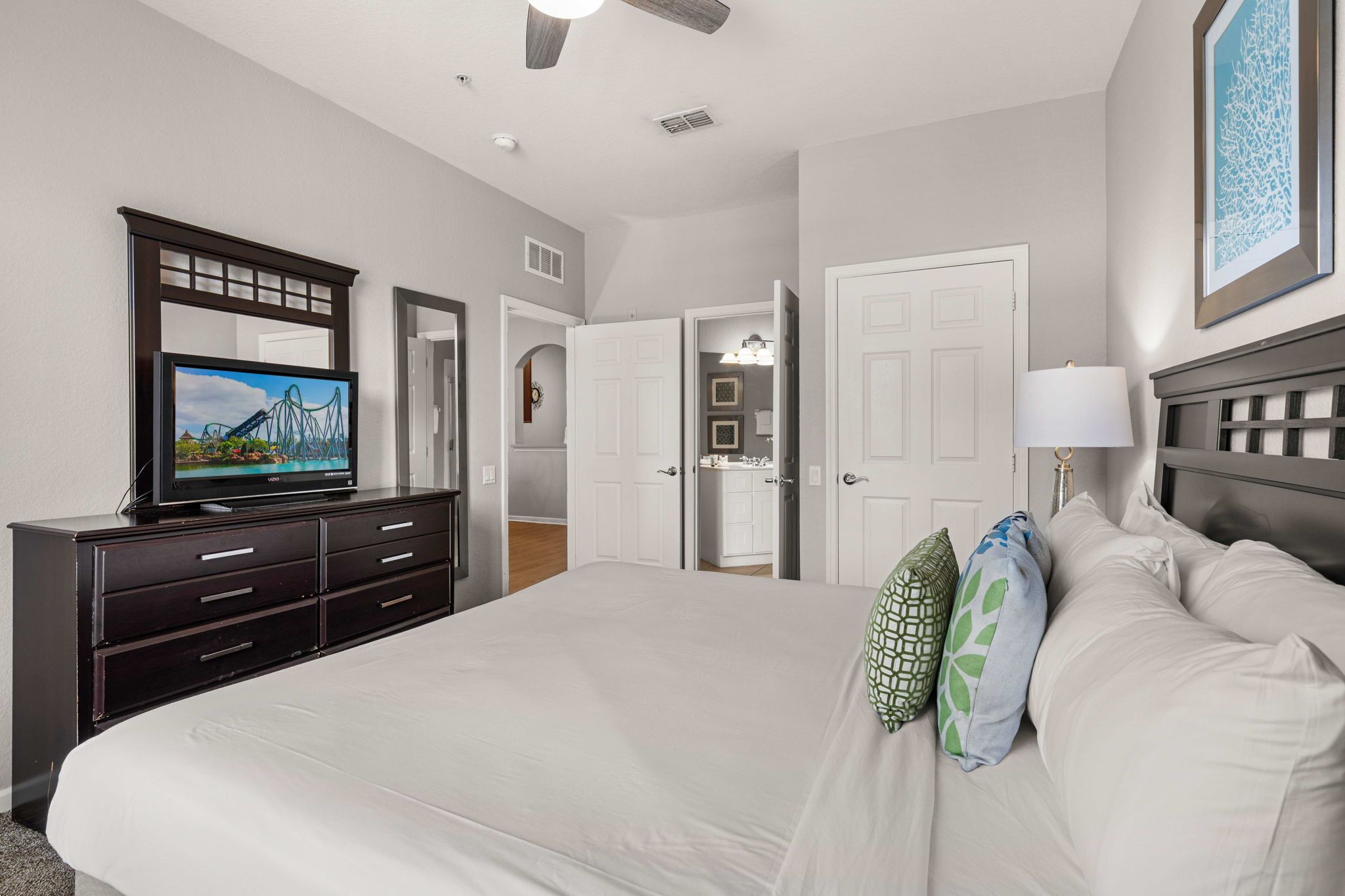 A hotel room with a large bed, a TV on a dresser, a door leading to a bathroom, and a ceiling fan.