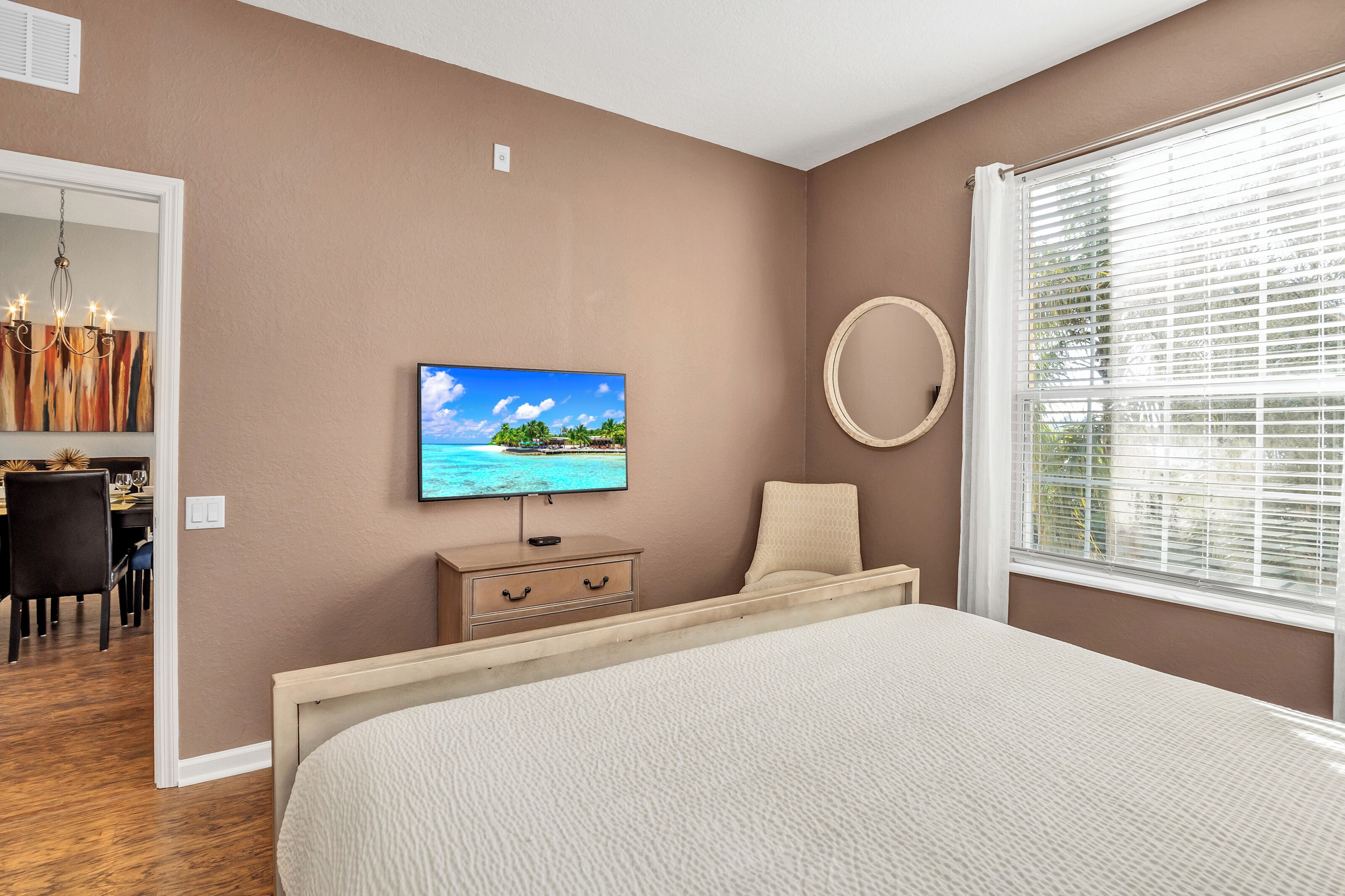 Une chambre à coucher comprenant un lit, un téléviseur fixé au mur, un bureau avec une chaise et une fenêtre avec des stores.