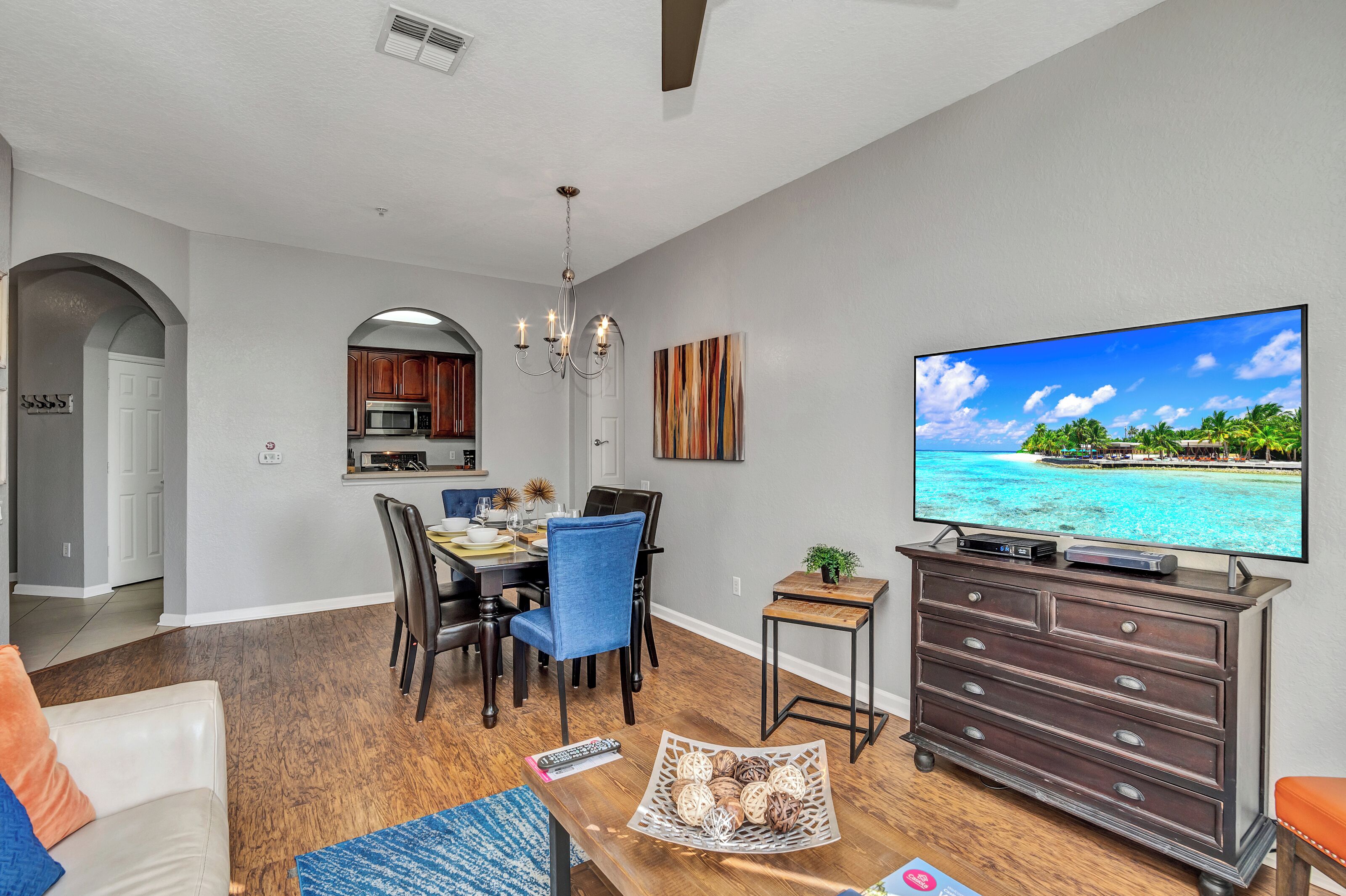A modern living room with a large flat-screen TV, a dining area with a chandelier, and a kitchen visible through an archway.