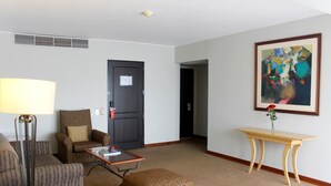 A hotel room with a sofa, armchair, ottoman, a small table, a lamp, a painting, and a door. - El Sol De Oro Hotel (Ollantaytambo)
