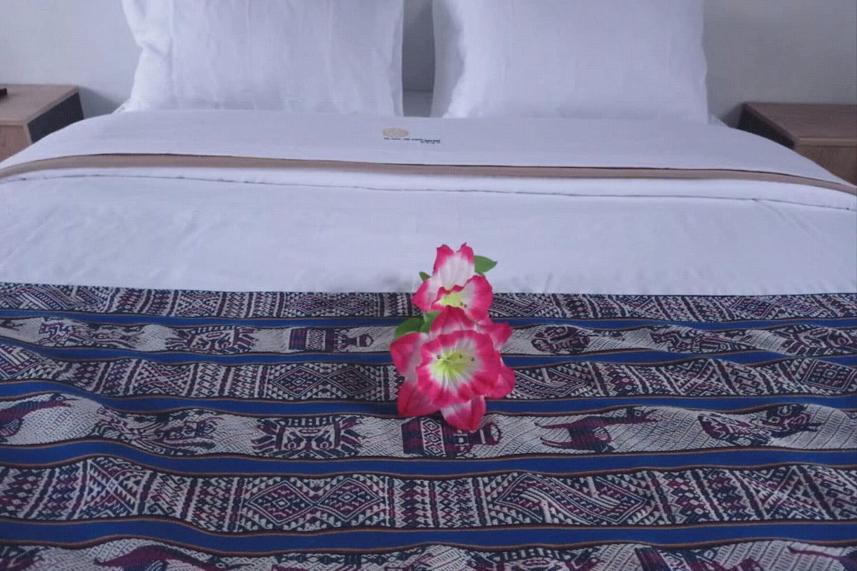 Un lit avec des draps blancs, une couvre-lit à motifs et une seule fleur rose.