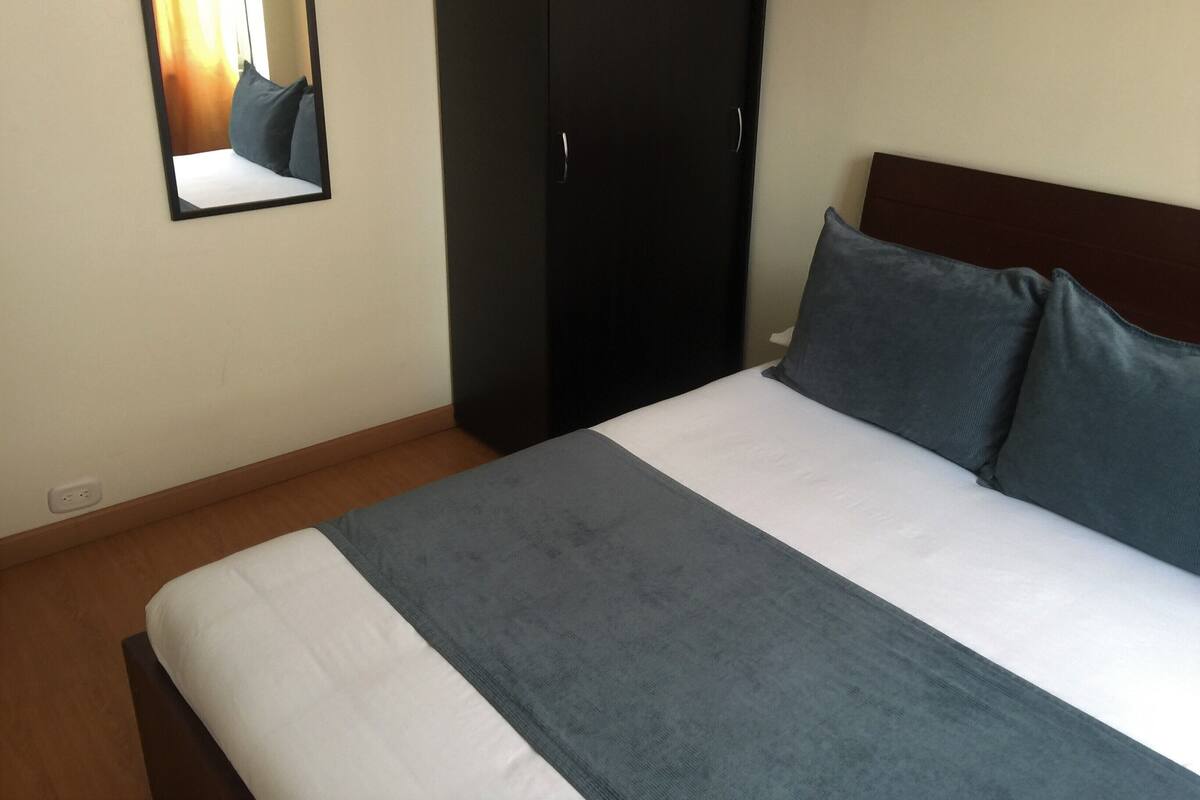 A neatly made bed with a white and gray bedspread and two pillows. A dark wardrobe is against the wall, and a mirror hangs above the bed.